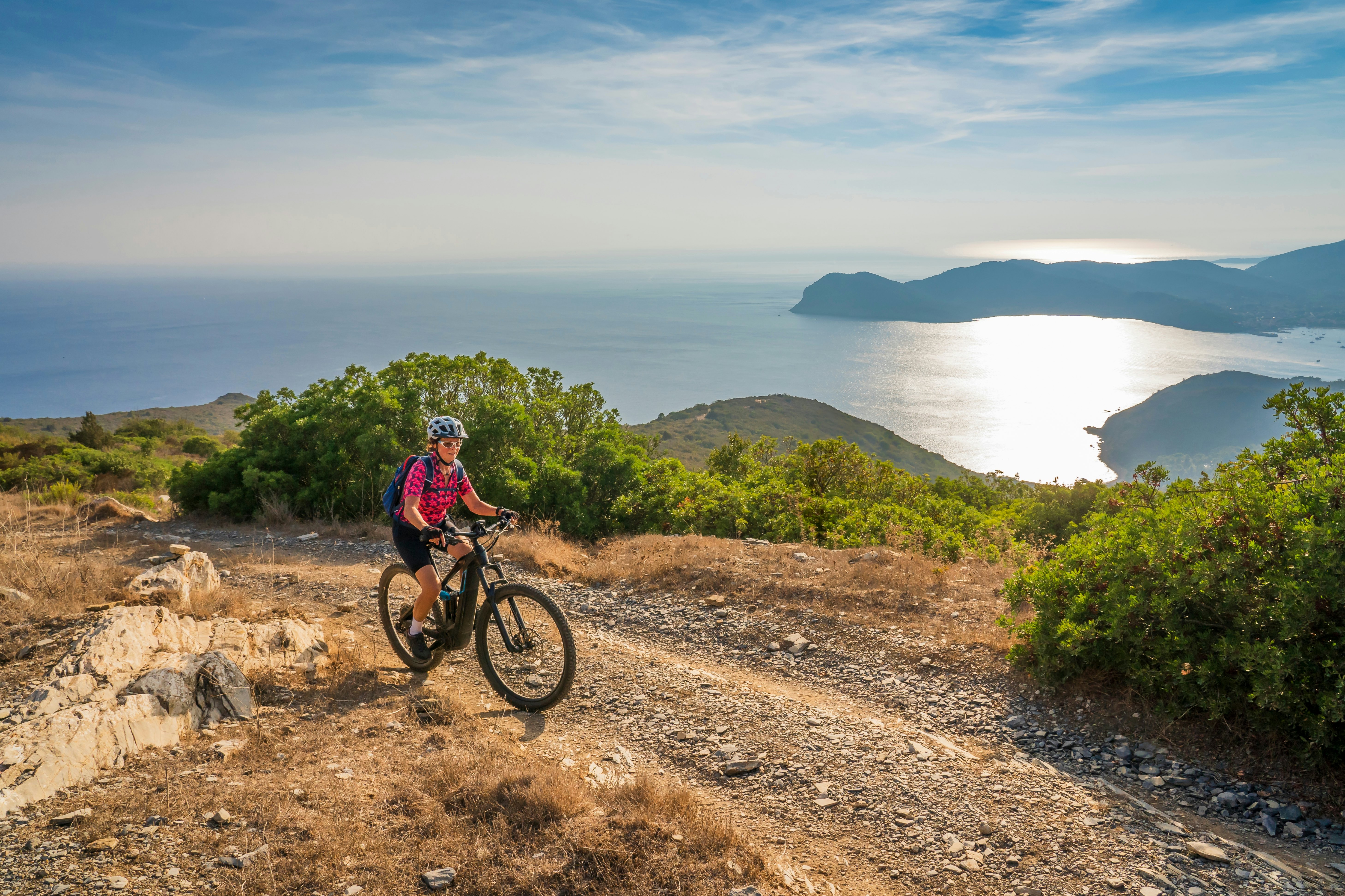 A woman rides an e-bicycle along a dirt trail on a hill overlooking the sea.
