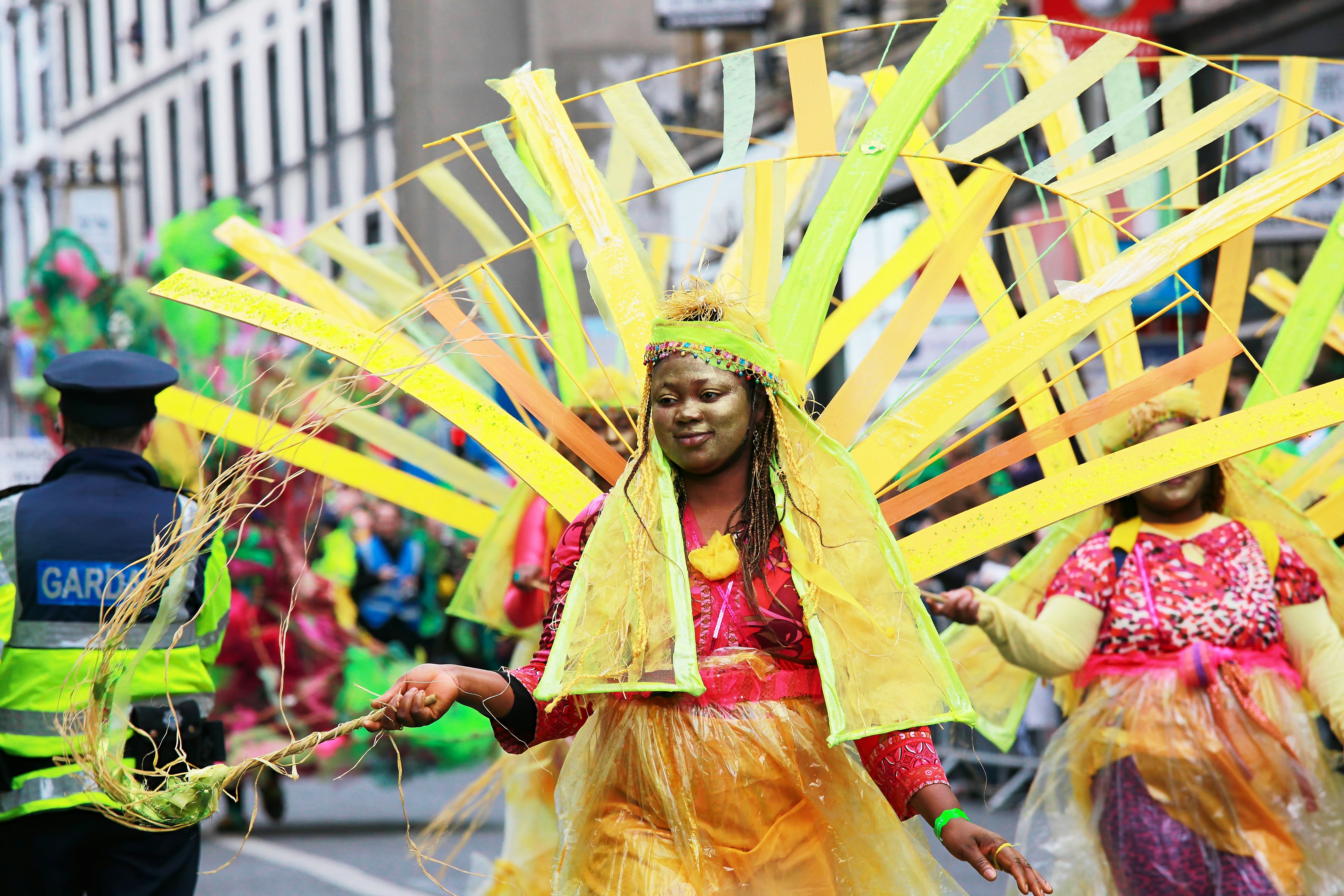 Women in pink tops and yellow skirts with large yellow headdresses wave wands as they walk in a parade