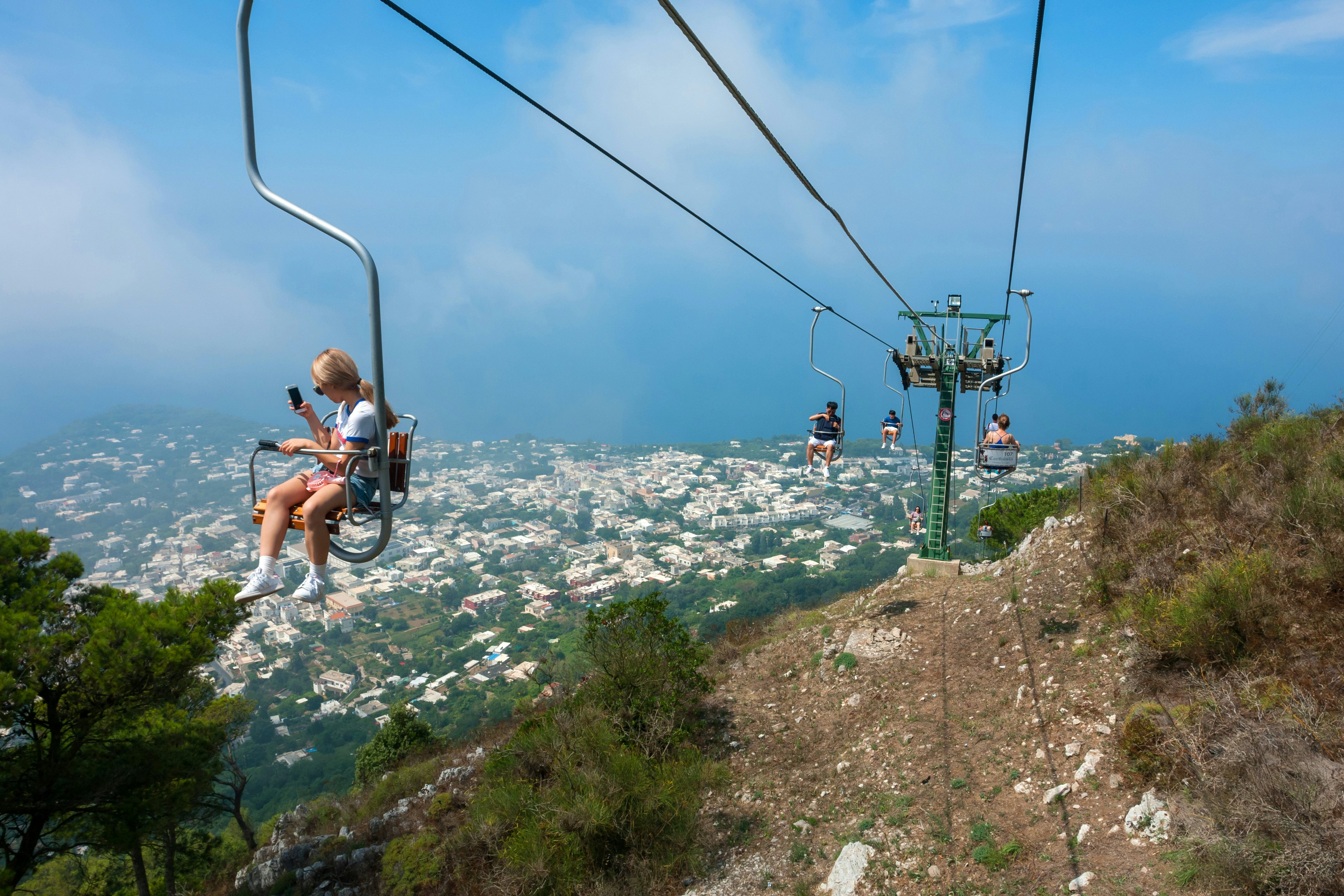 Tourists riding up the mountain chairlift on the island of Capri, Italy, with a view of houses and the coast behind.