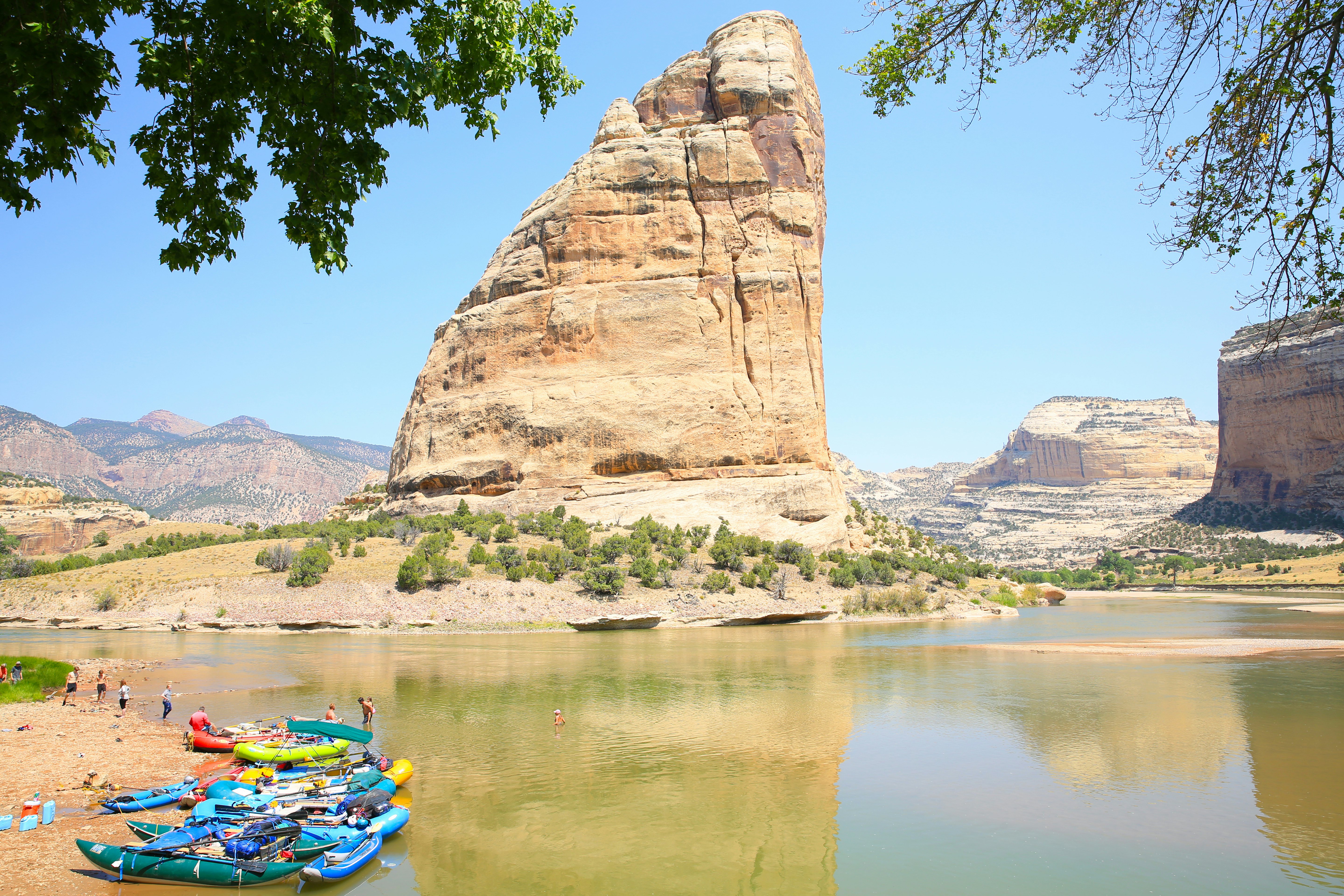 Kayakers at the shore of a river bend around a vast sail-shaped rock.