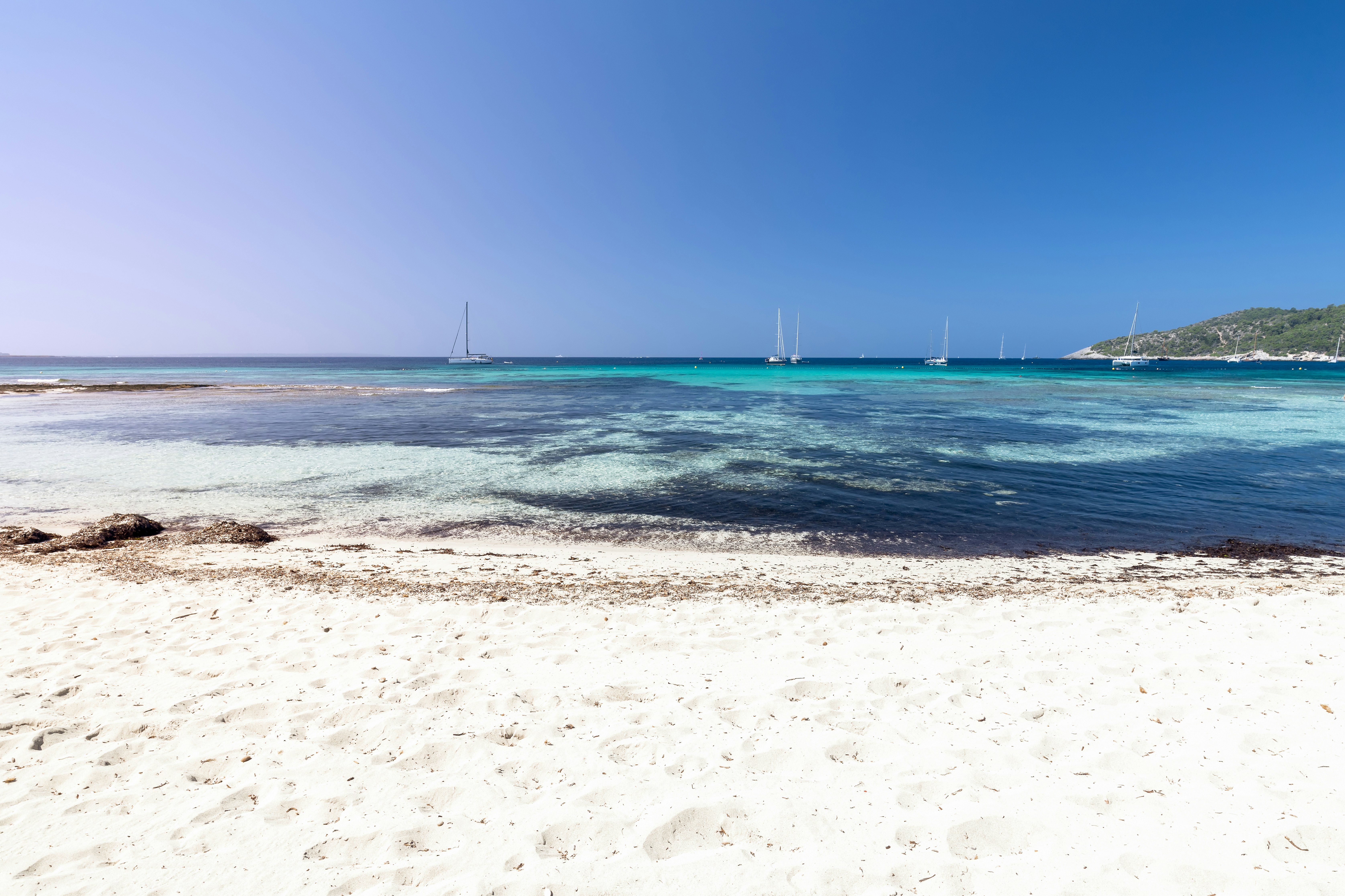 A view of an empty white-sand beach. Sailboats are visible in the azure waters of a cove beyond the beach.