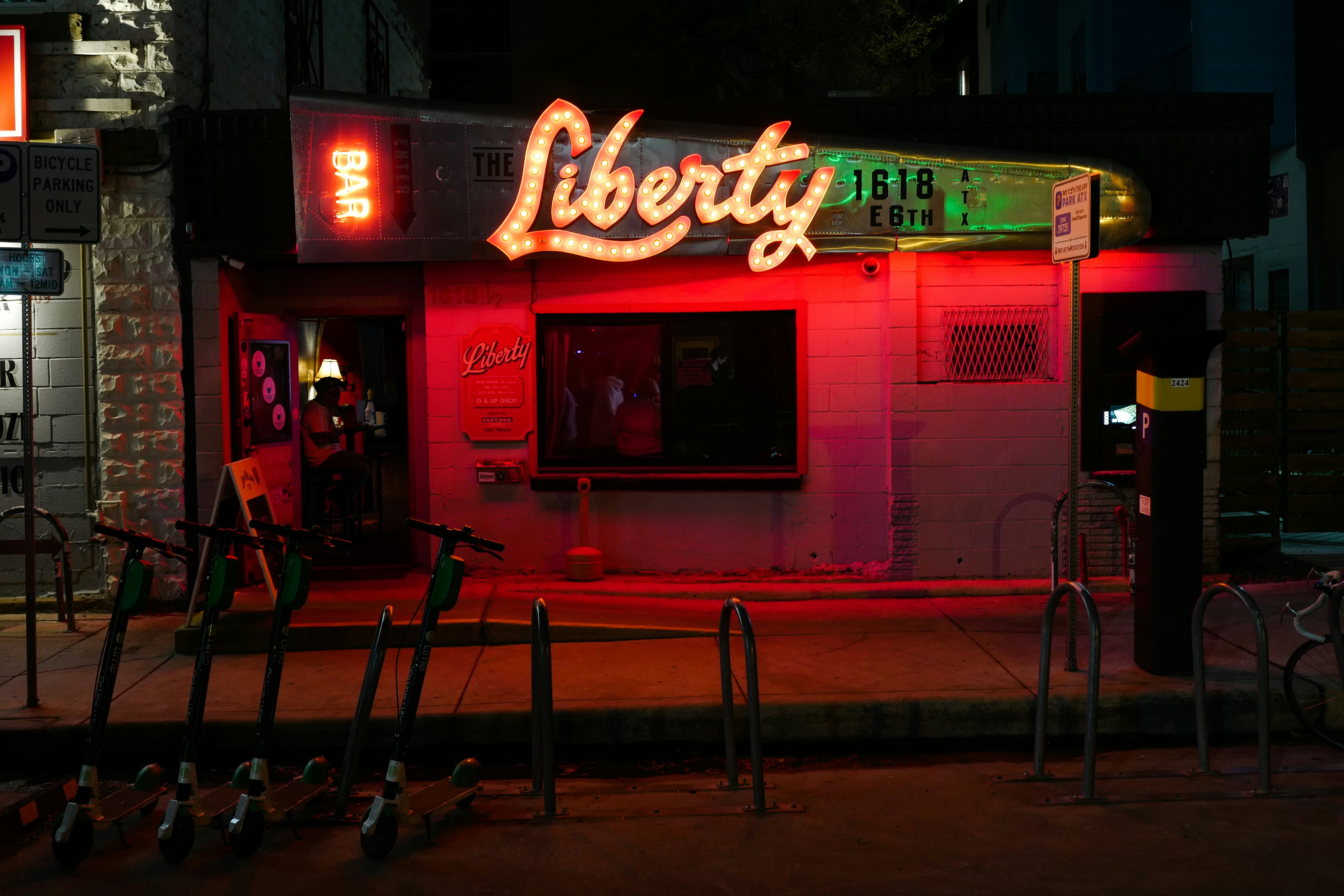 The facade of a dive bar on a city street is illuminated by red lights and light-bulb letters.