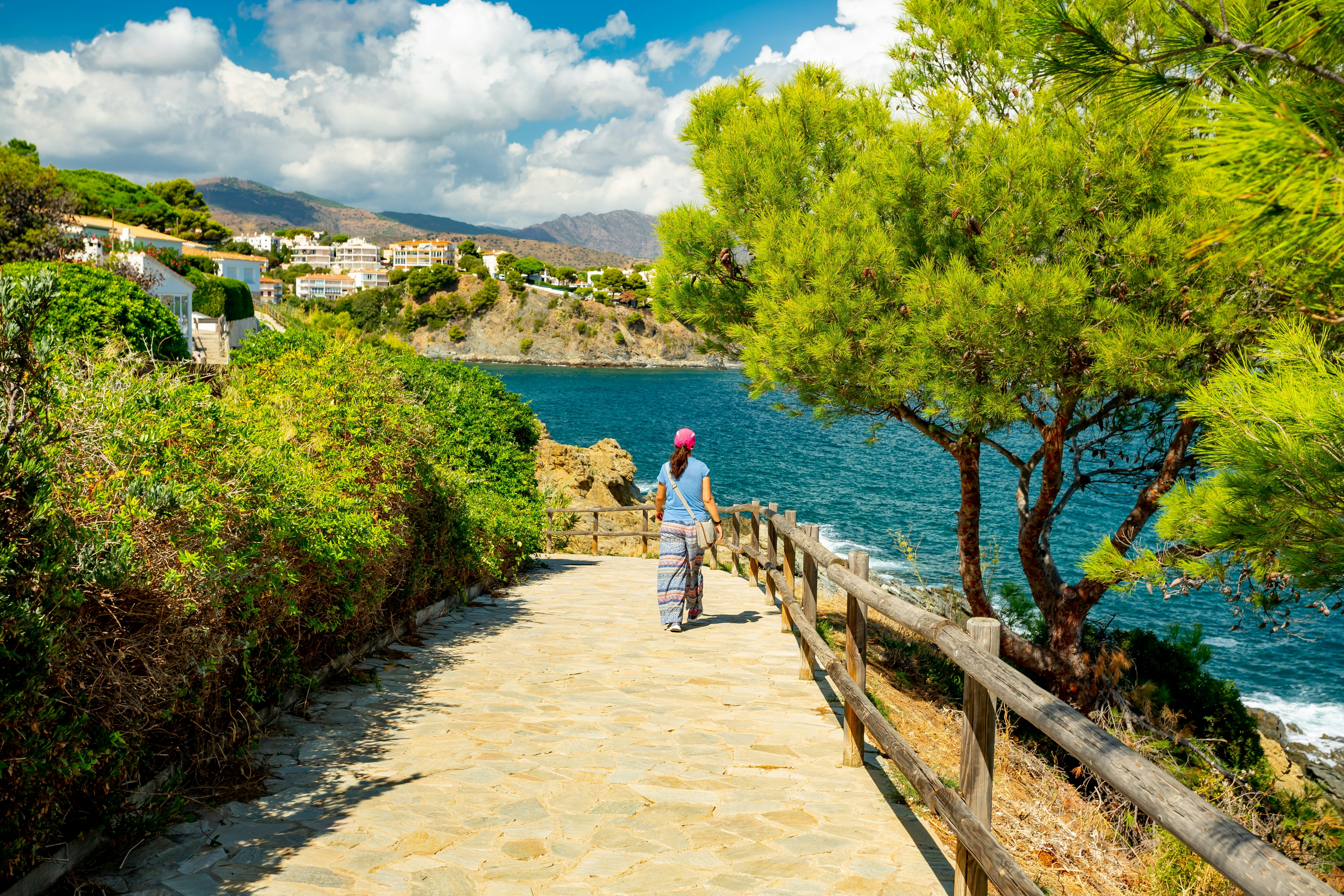 A woman in loose-fitting pants is seen from behind walking on a paved path next to the sea, with vegetation lining the trail.