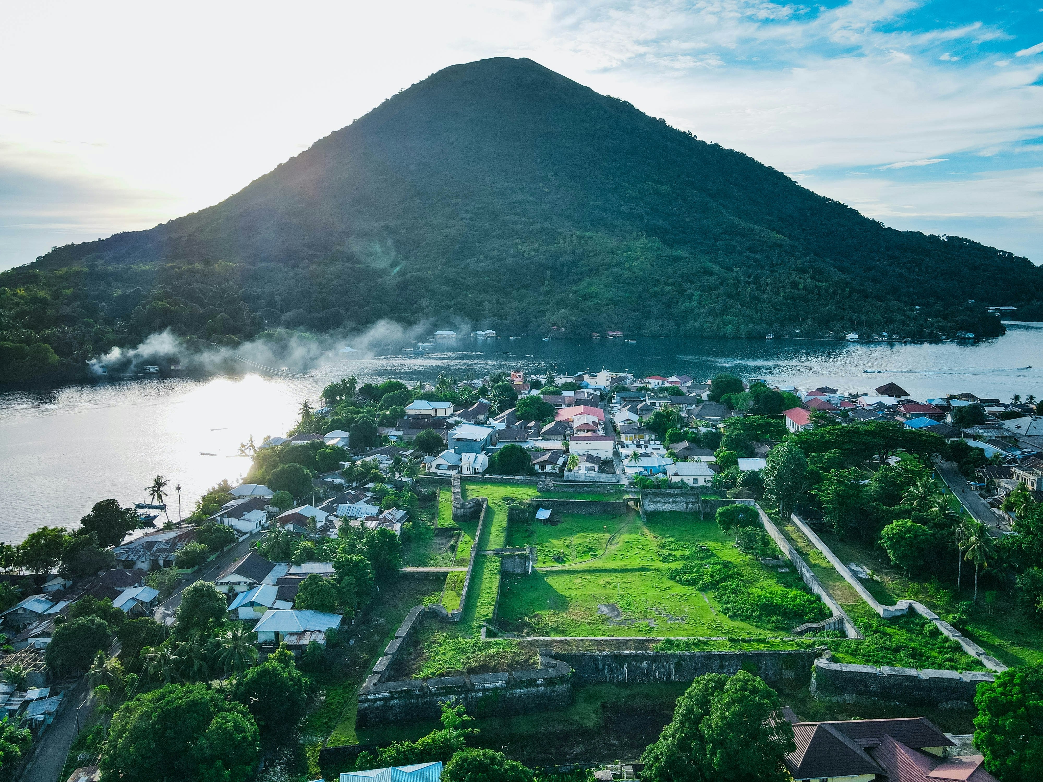 An aerial view of the outline of a former fortress now covered in grass, next to a village and beside a channel between hilly green islands.