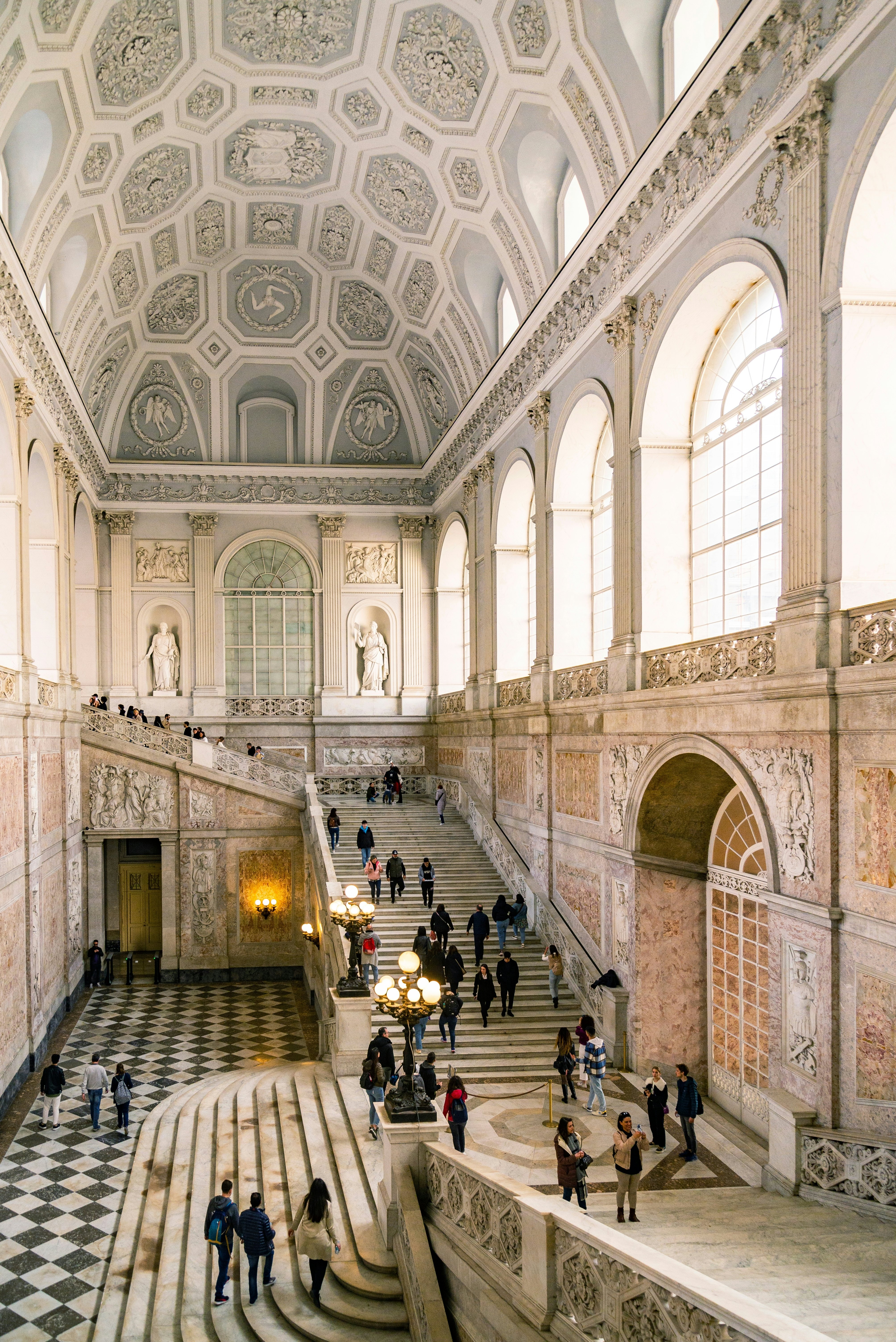 A grand entrance hallway with marble floors and stairs leading upwards to the second floor. The ceiling is covered in relief artworks.