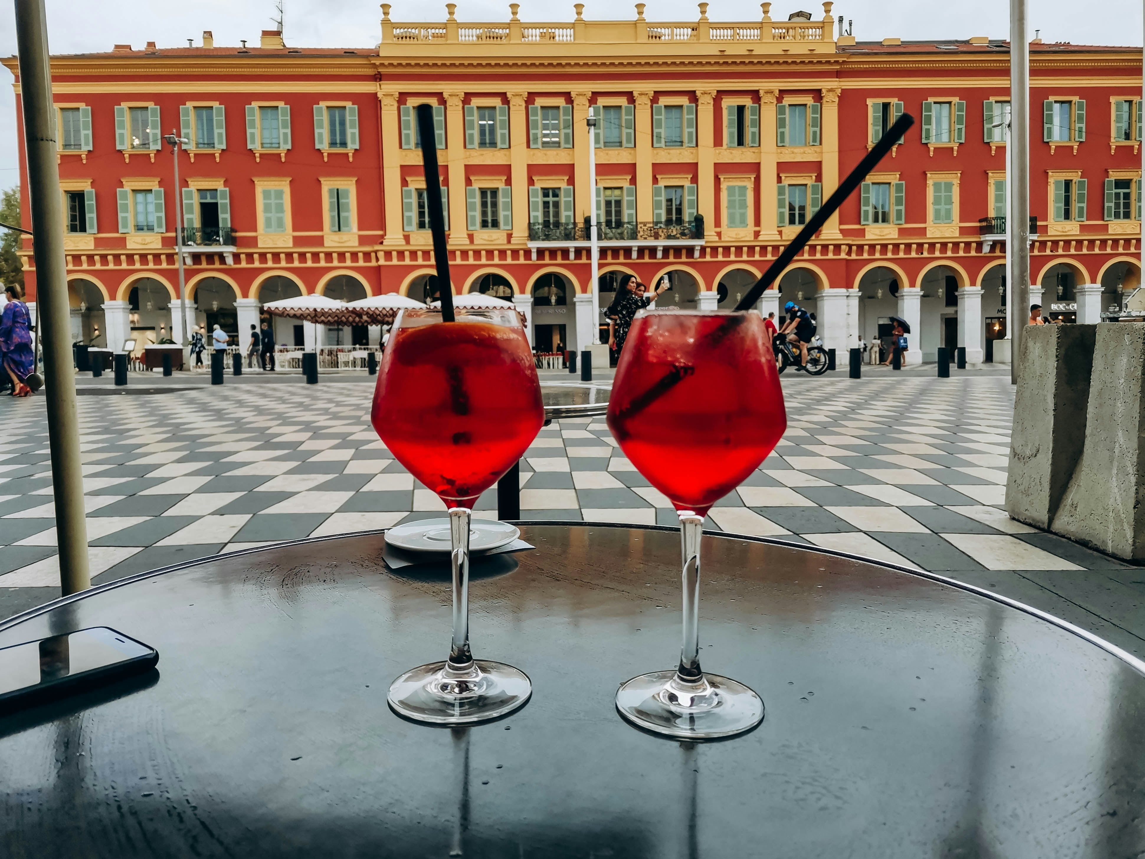 Two pink-colored cocktails sit on a table next to a city square with checkerboard pavers, and a colonnaded palace on the far side.