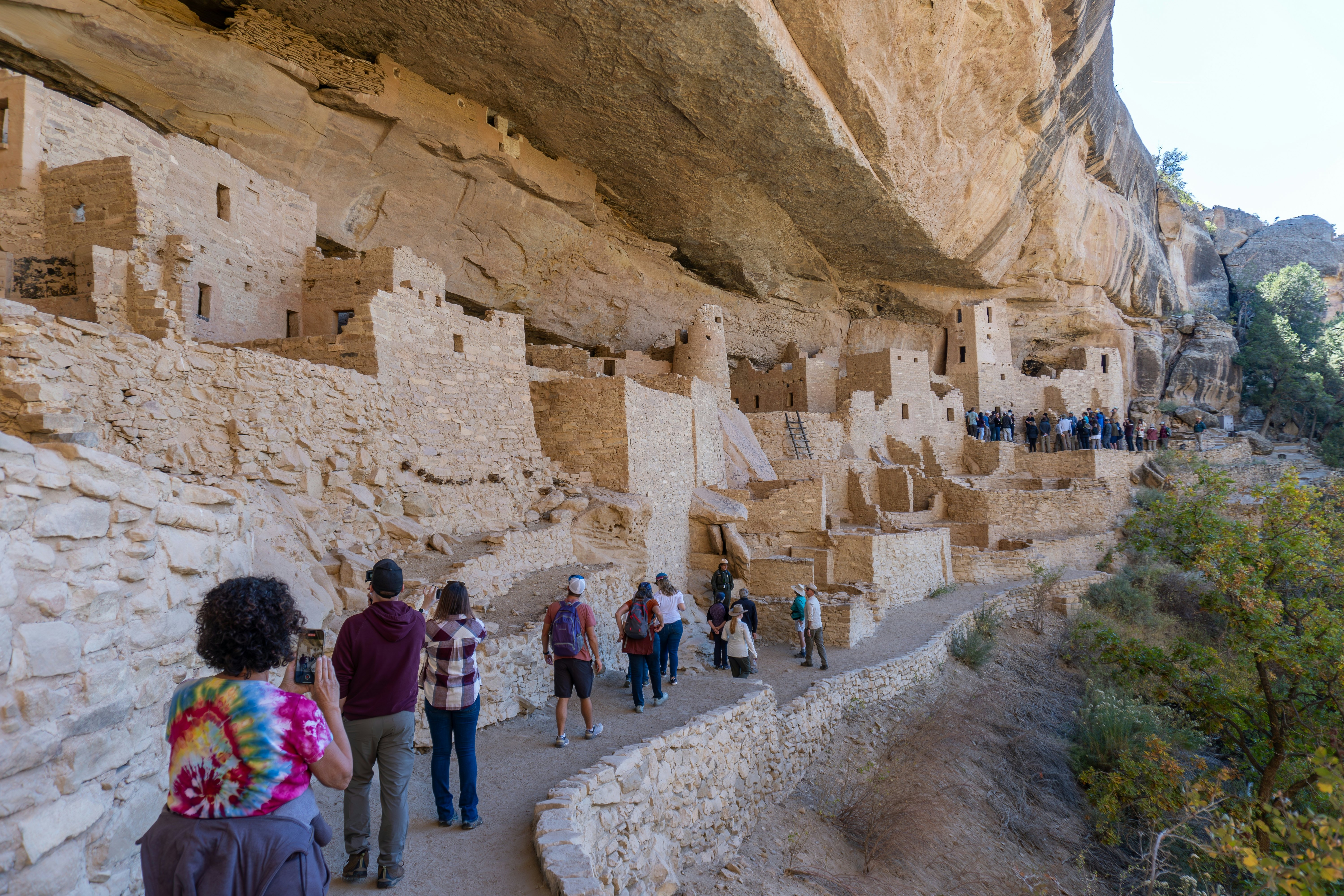 Rangers lead tourists along a path beside ancient brick dwellings built under the overhang of a cliff.