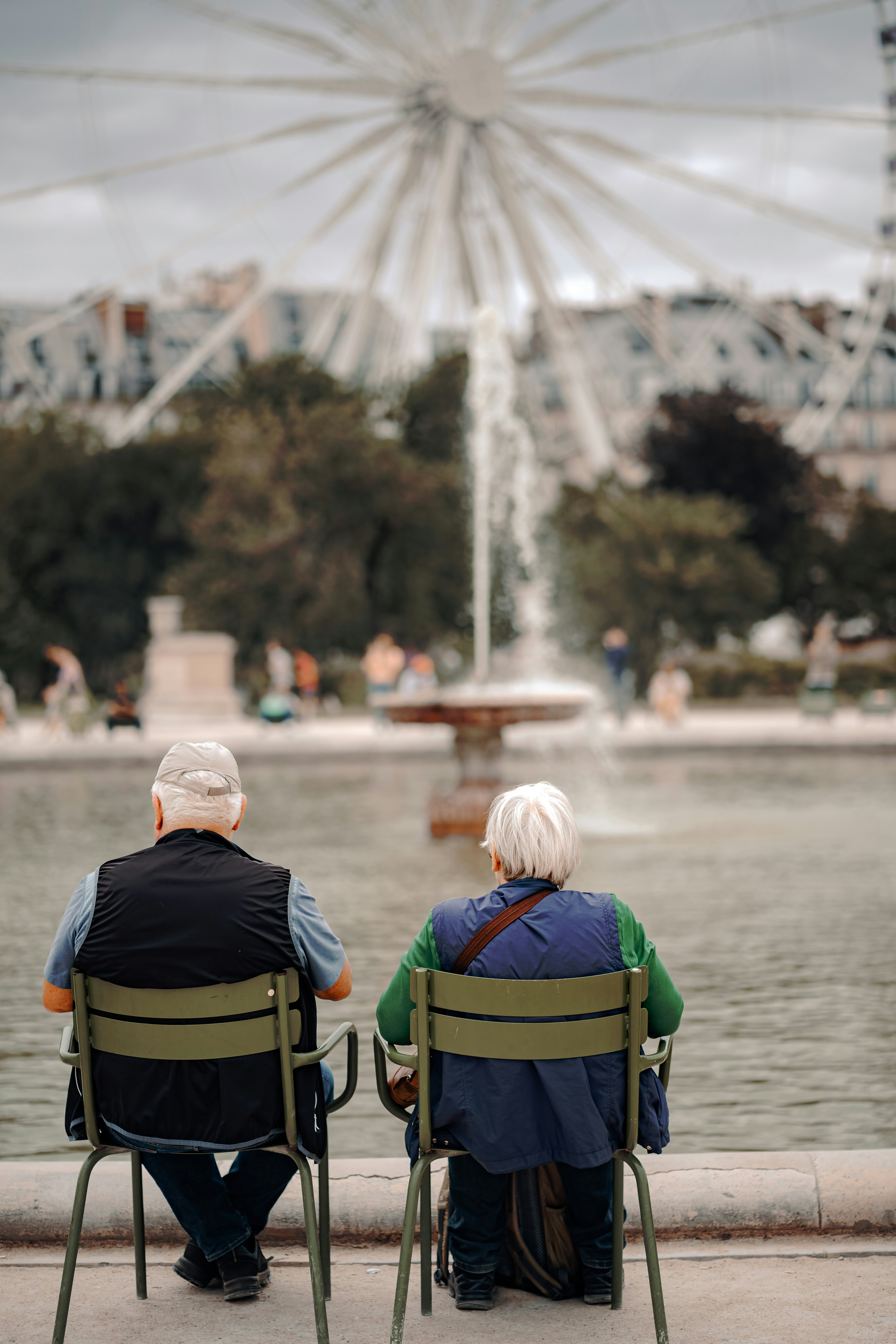Paris, France - 13.07.2023: an elderly couple sits on green chairs next to a public spring fountain in a park in Paris,