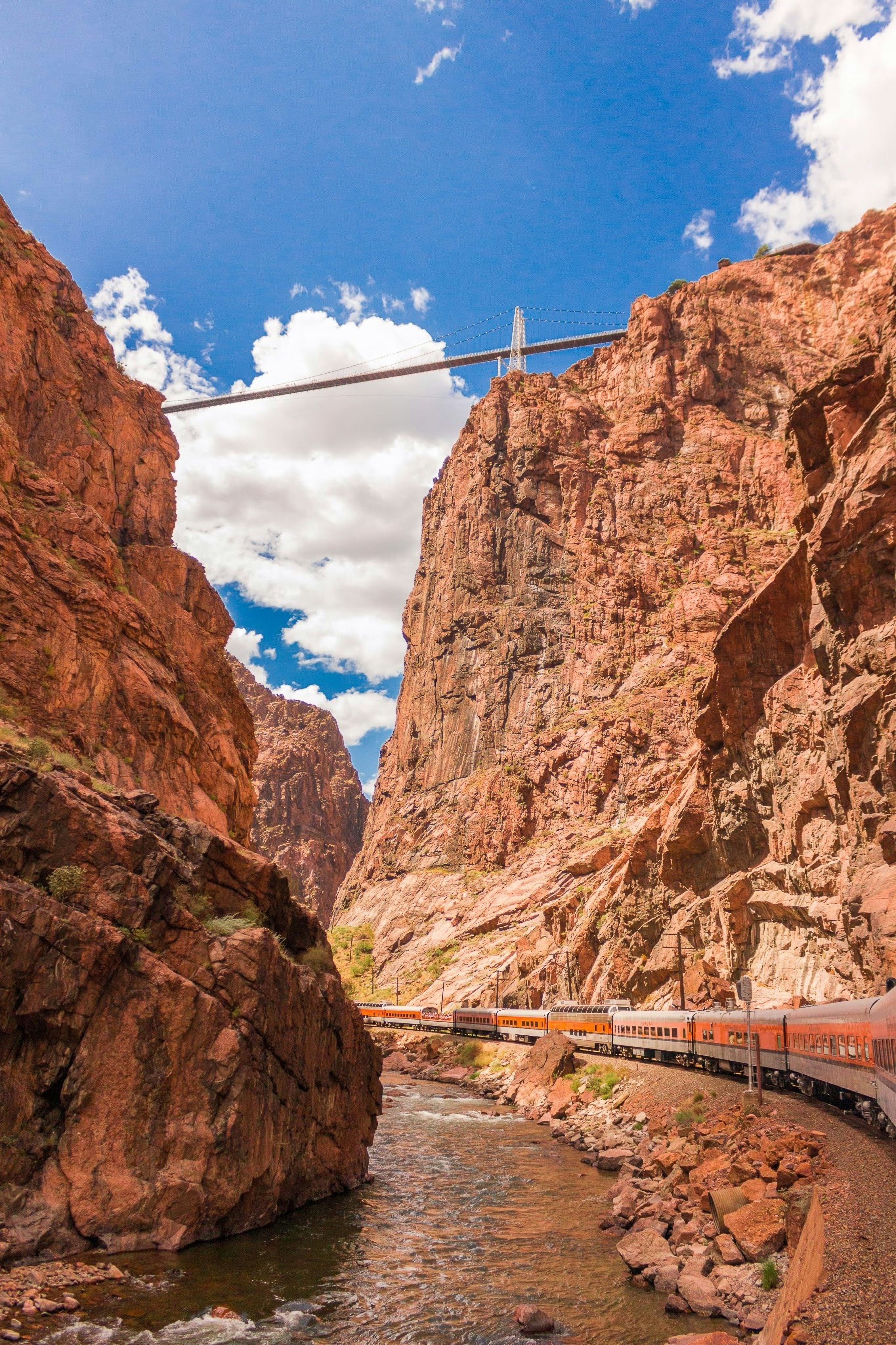 A train passes through a river gorge with steep red rock sides. High above is a suspension bridge.
