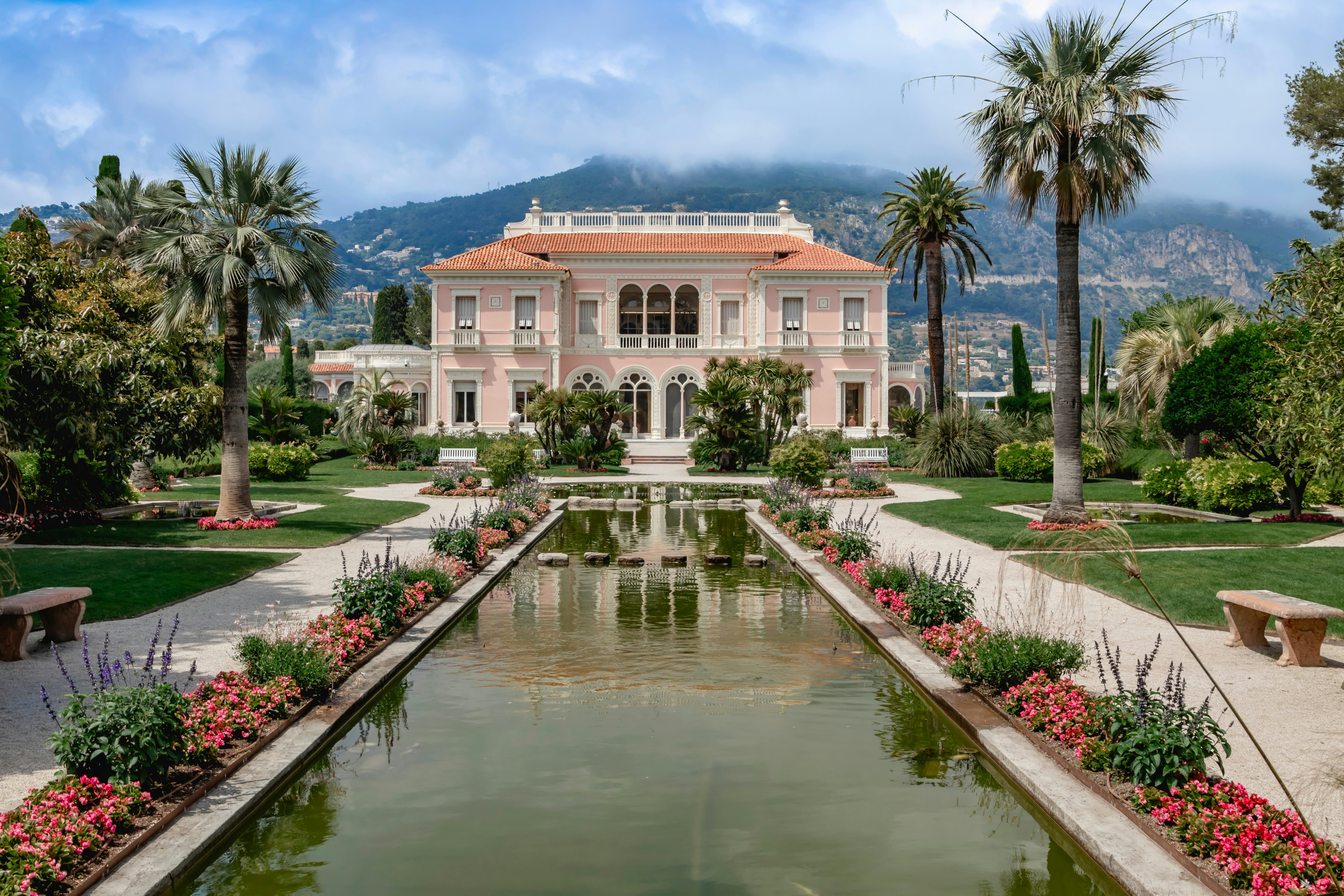A pink-painted villa sits among beautiful gardens and a tranquil water feature set against scenic mountains.