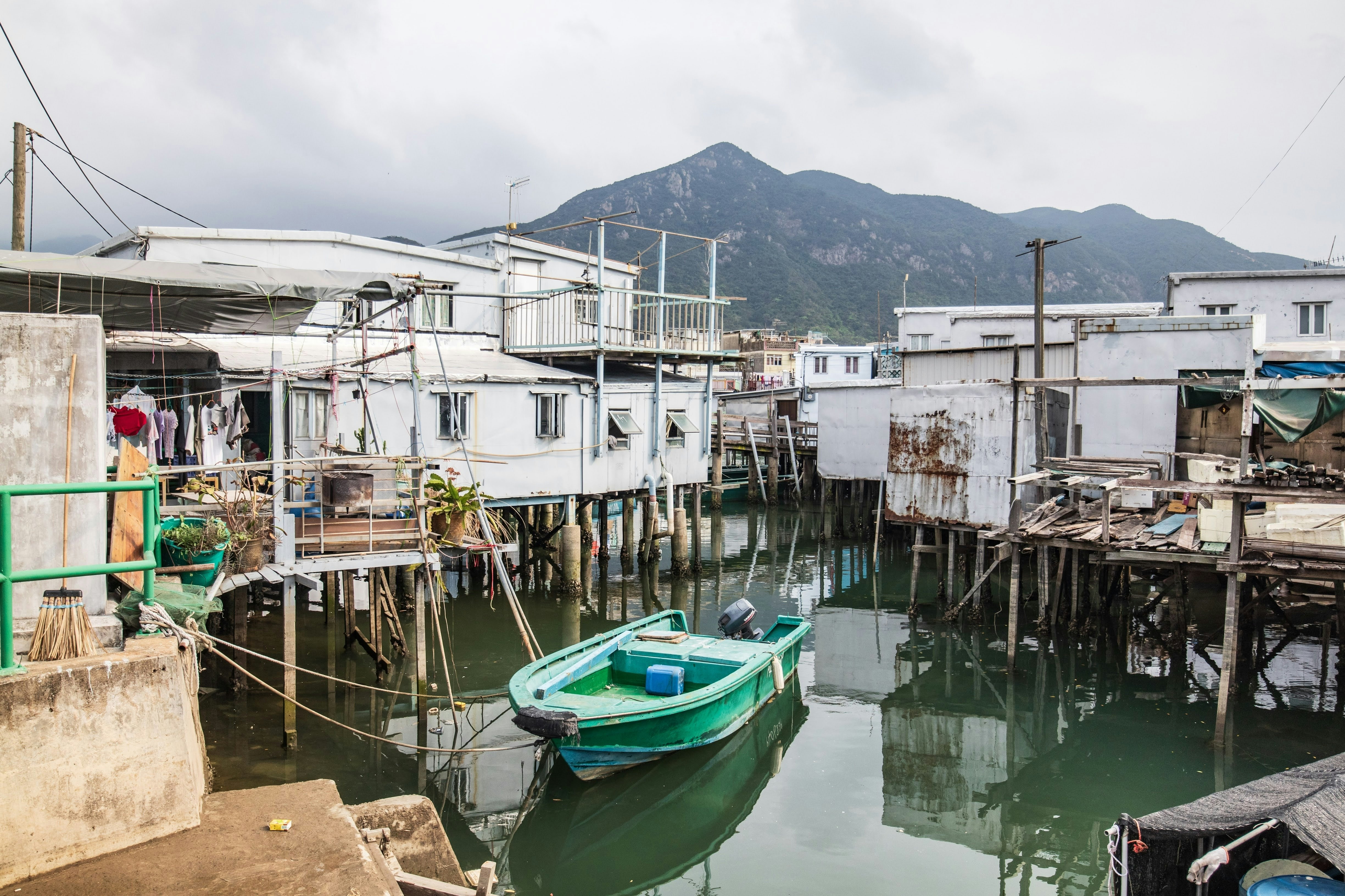 A green boat is moored in a small channel surrounded by houses on stilts in the water.