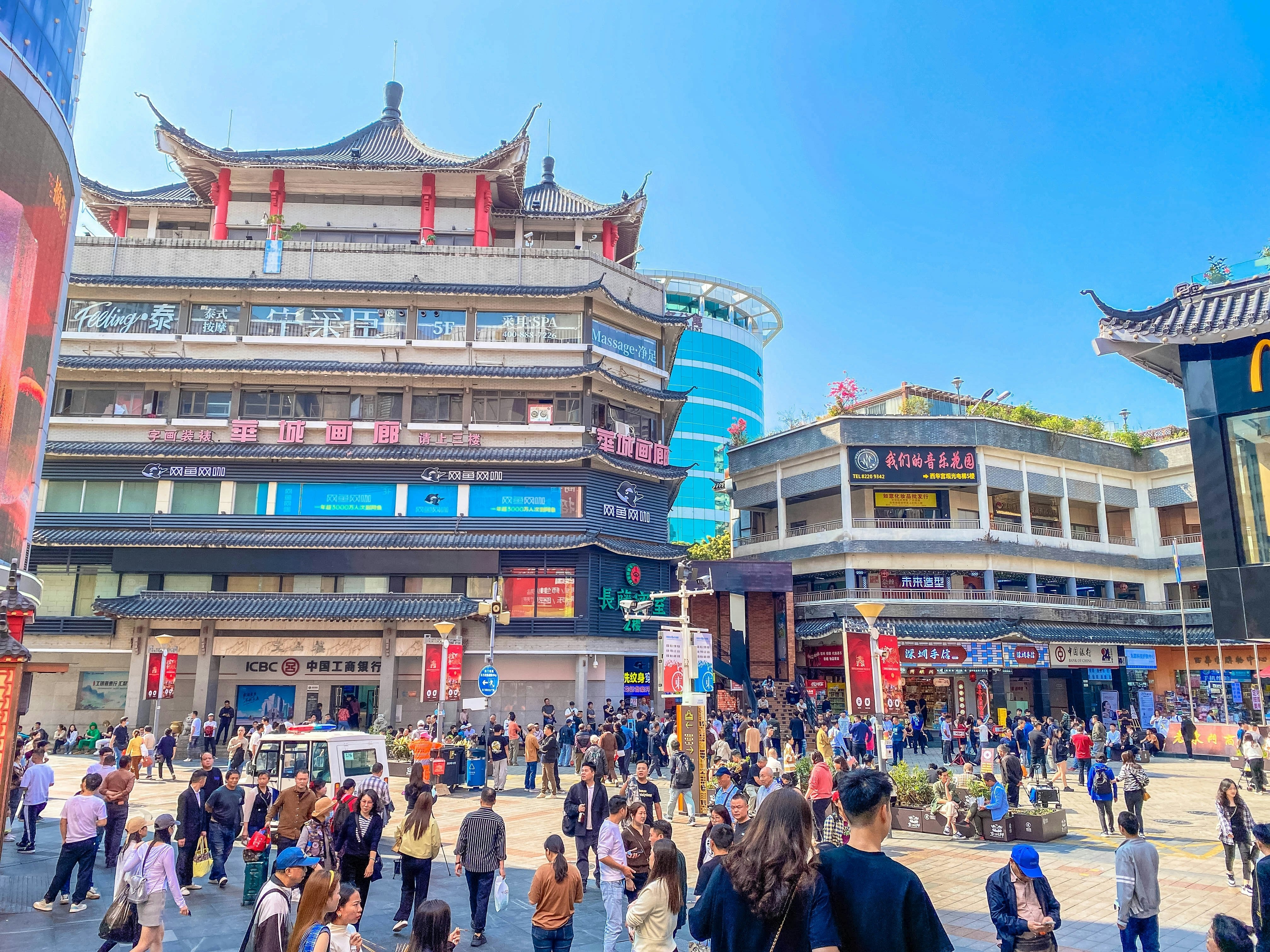 People crowd a city plaza surrounded by buildings with shops and restaurants.