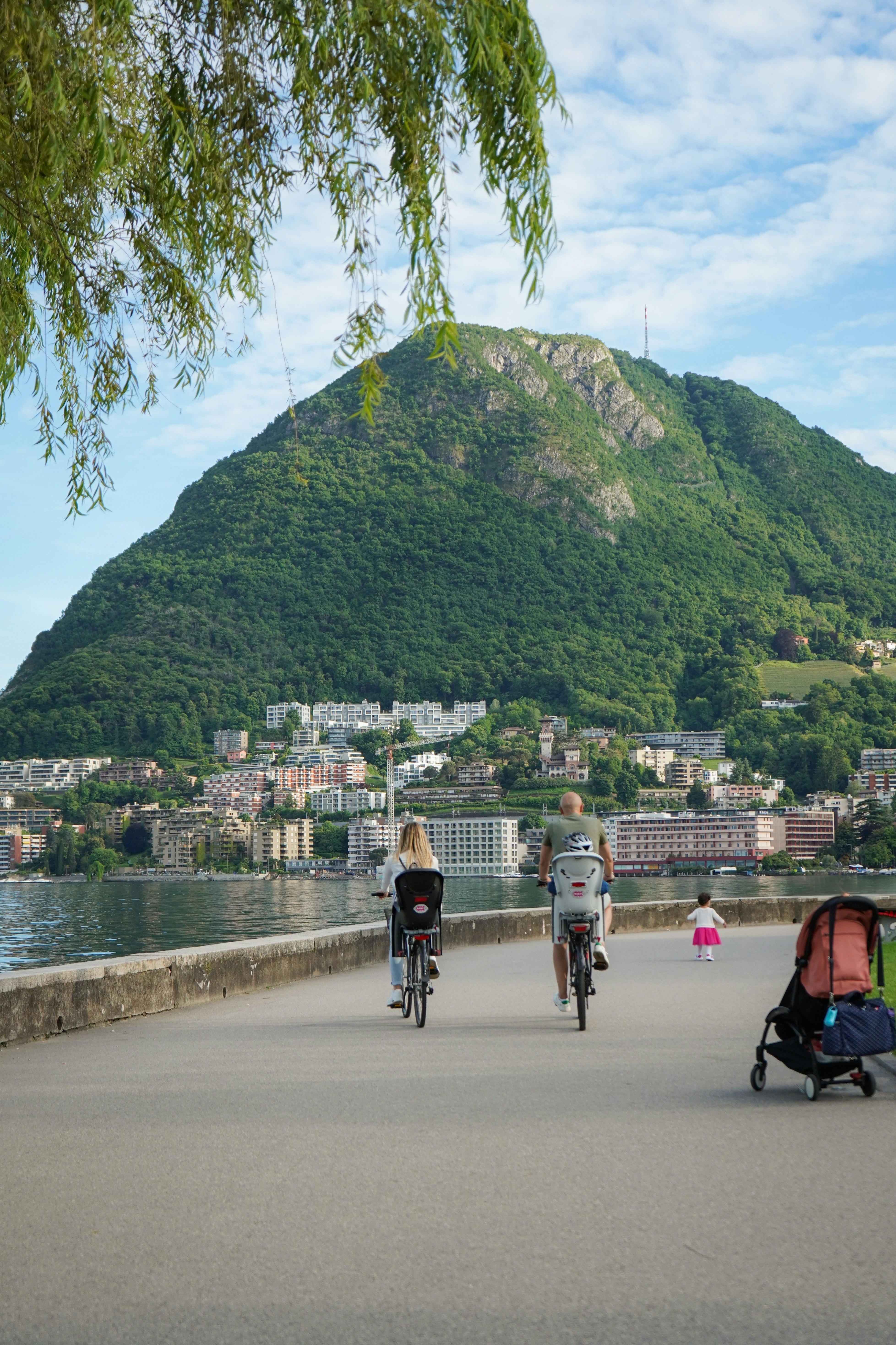 A couple cycling on bikes with a baby on the back and a small child running nearby beside a lake.