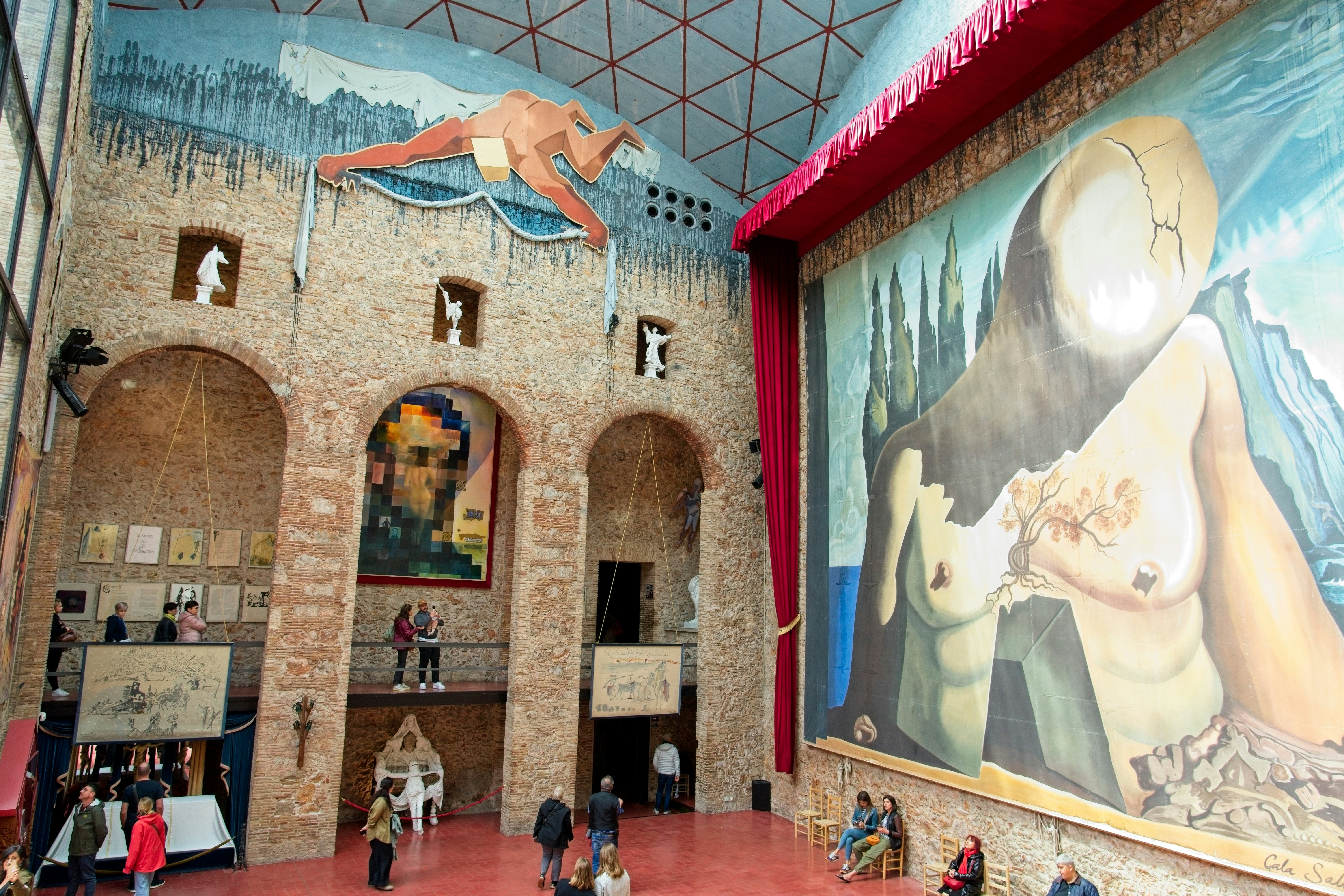 People walk in the interior of a public space displaying huge works of art, including a wall-size mural.