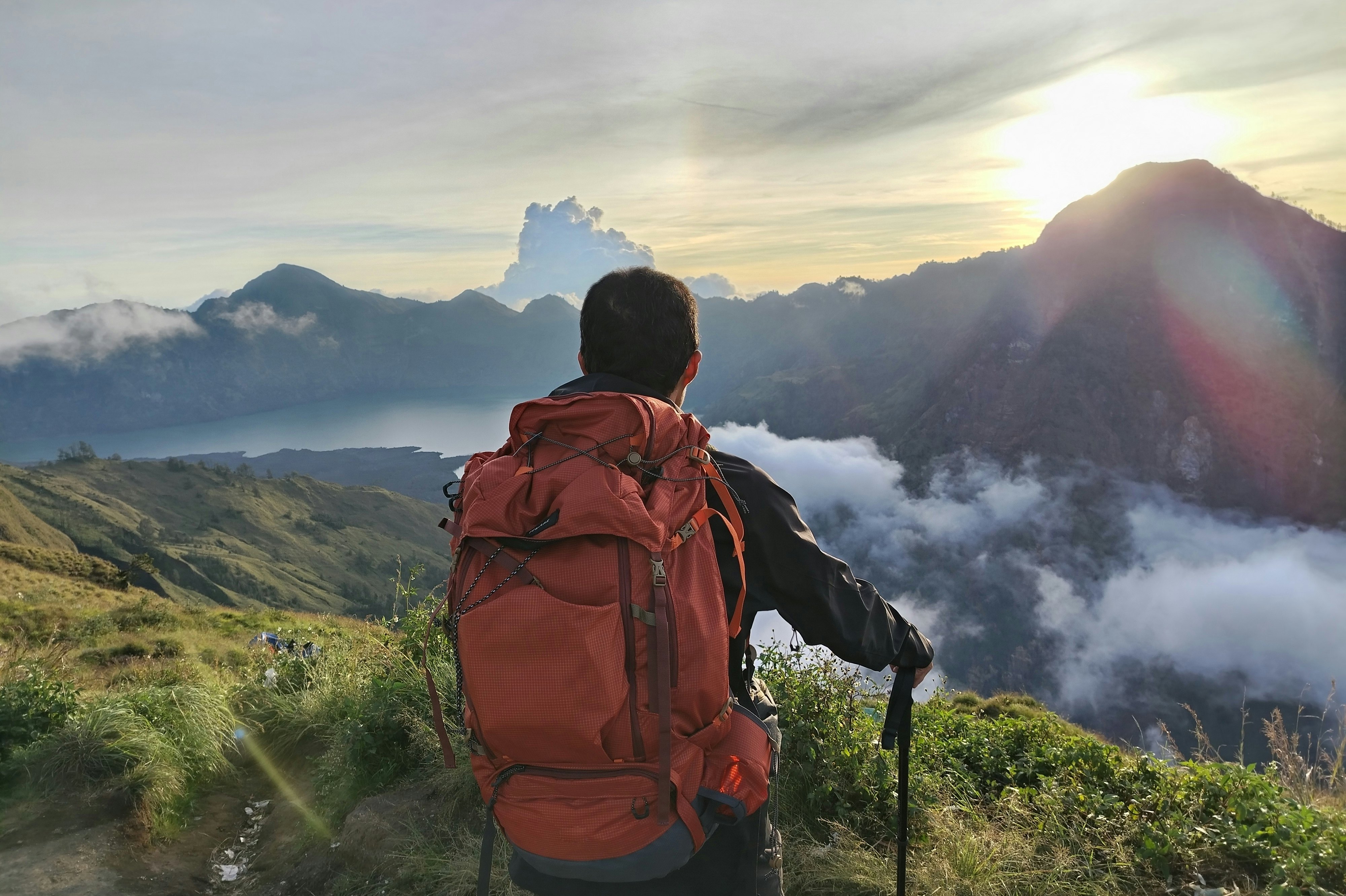 A hiker with a large pack is seen from behind on a grass-covered ridge looking out at misty mountains at sunrise.
