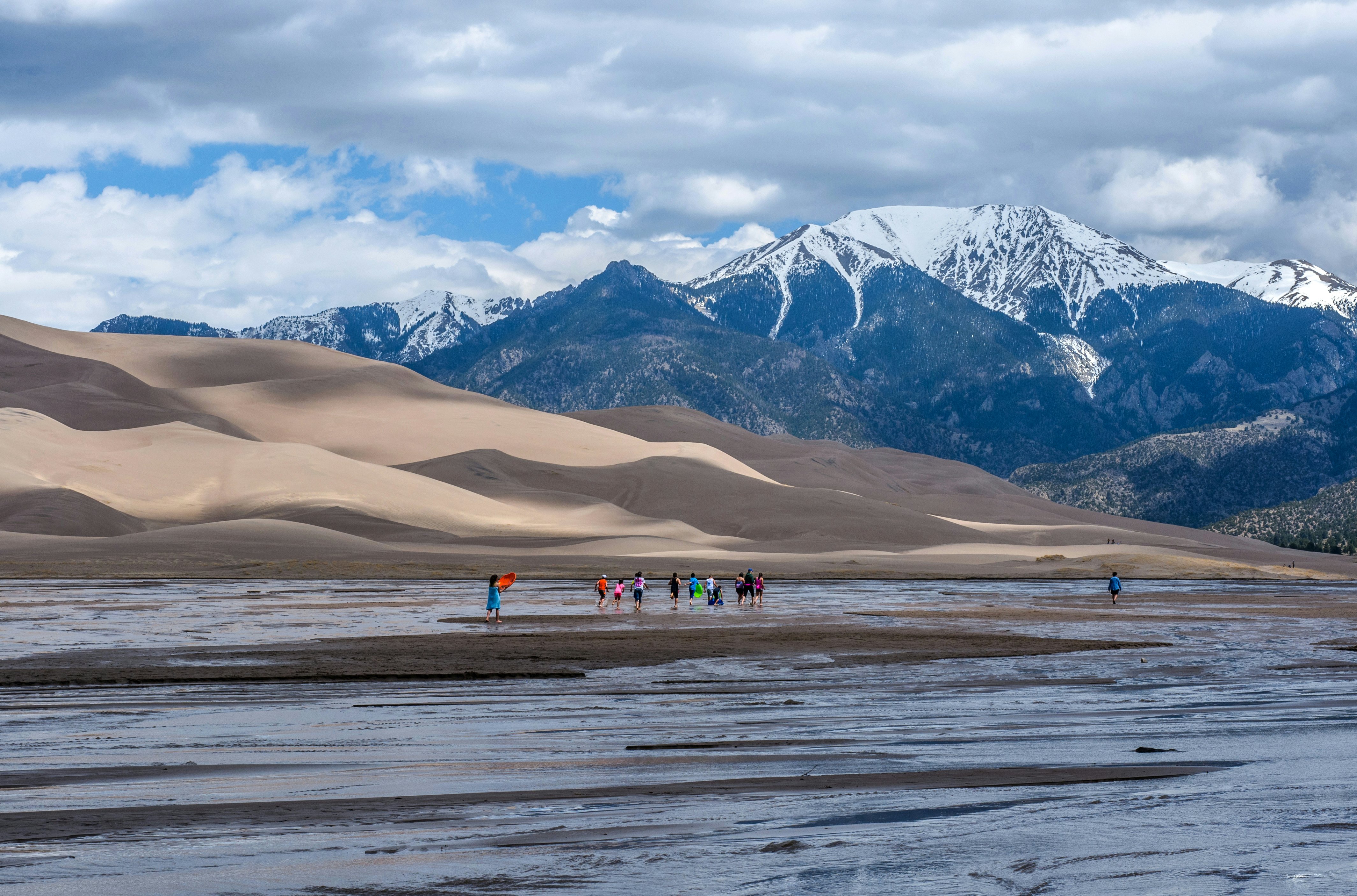 Great Sand Dunes National Park & Preserve, Colorado, USA. A group kids are playing in Medano Creek at the base of sand dunes and snow-capped peaks as spring clouds passing over.