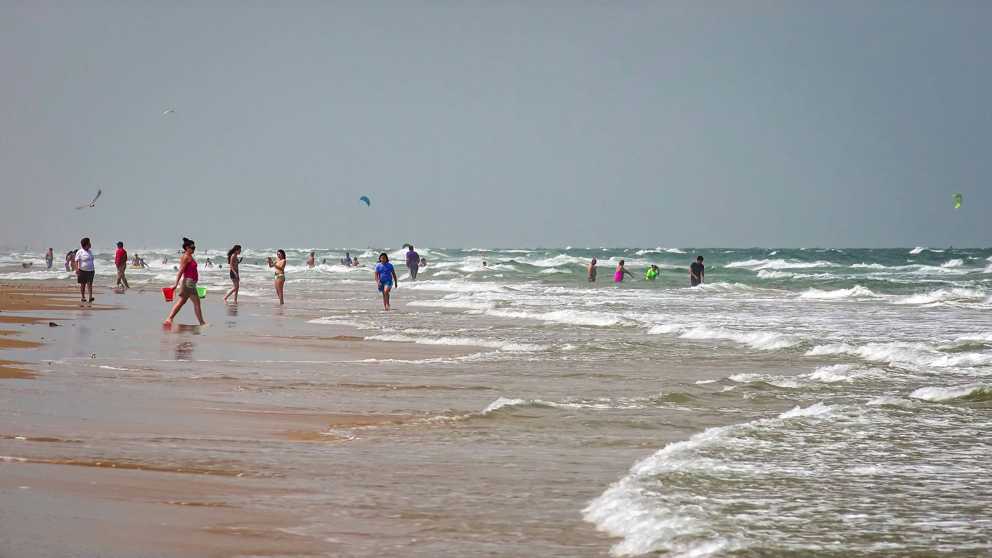 People wade into the white-capped waves that lap a wide beach.