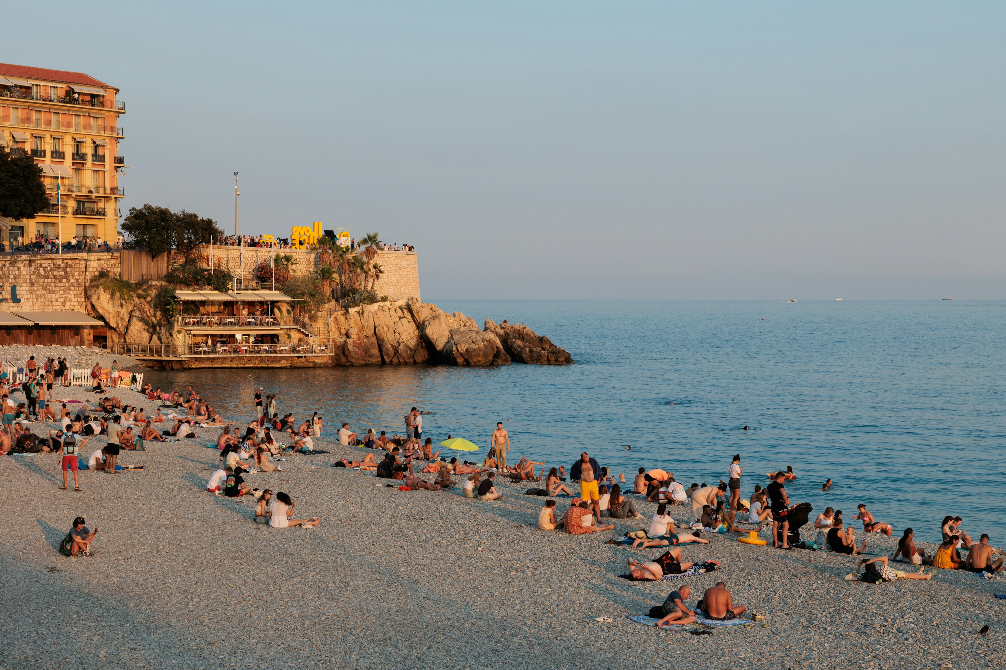 People sit on a pebbled city beach late in the day. Light hits buildings along the shore beyond the beach.