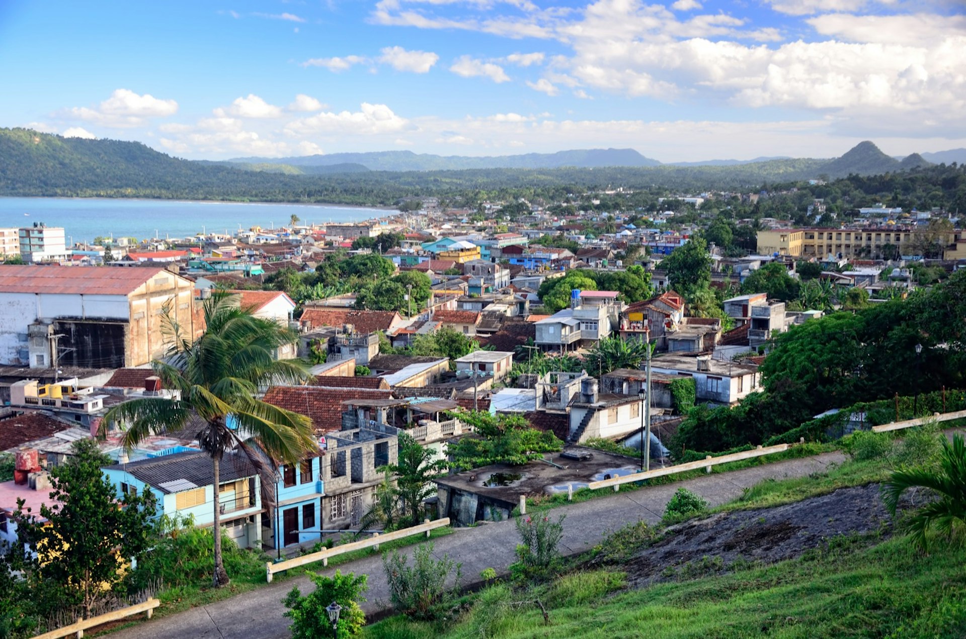 An aerial view of the town of Baracoa in Cuba. The Caribbean town is filled with red-roof buildings and palm trees. You can also see the winding paved road. 