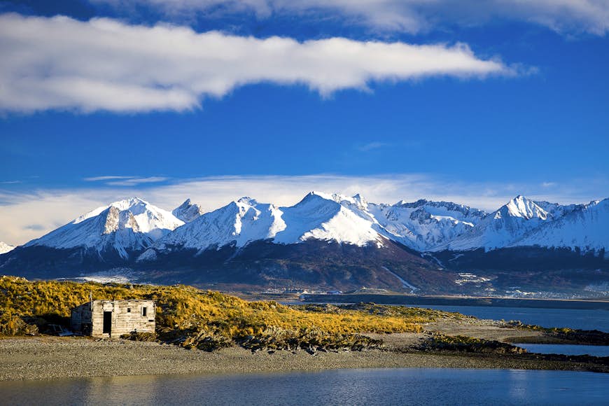 En landskapsvy av en liten byggnad mot en bakgrund av kustlinjen och snötäckta berg i Tierra del Fuego, Argentina