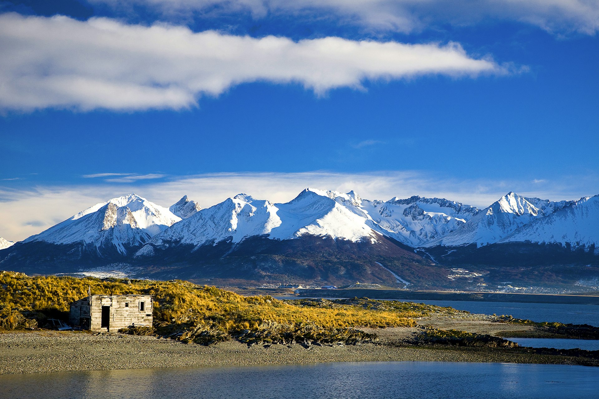 A landscape view of a small building against a backdrop of coastline and snowcapped mountains in Tierra del Fuego, Argentina