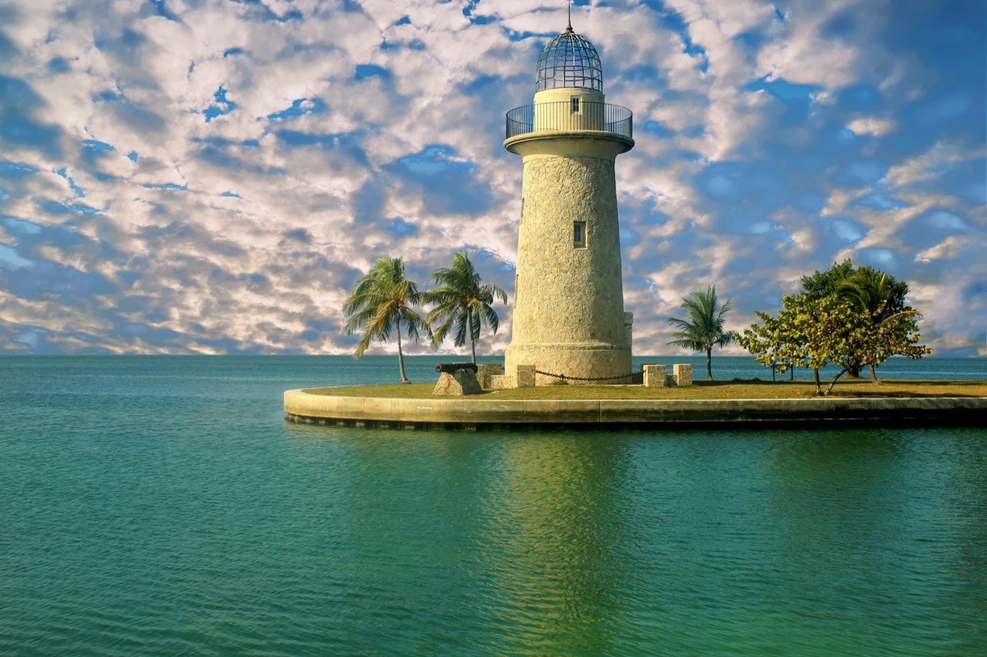 A conical lighthouse, with a few palm trees around its base, is framed by a cloud-filled sky on its spit of land jutting into a calm bay