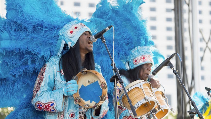 Two Mardi Gras indians in light blue feathered costumes play a tambourine and bongos on a stage; New Orleans Festivals