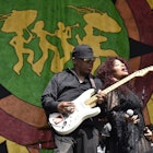 Disco legend Chaka Khan leans back against a man playing the electric guitar as she sings in front of a large tapestry; New Orleans Festivals