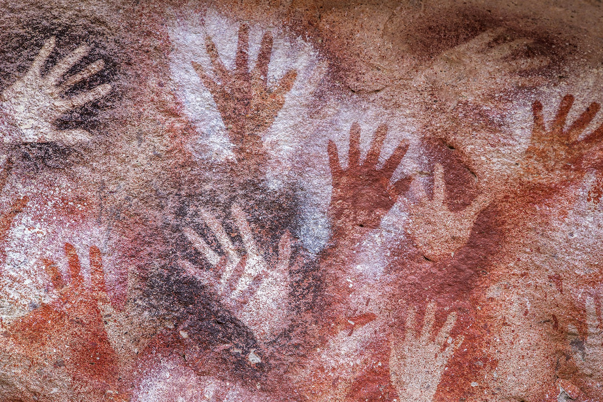 Hands made from painted shadows in white, red and maroon spread across an orange cave wall. Patagonia, Argentina.