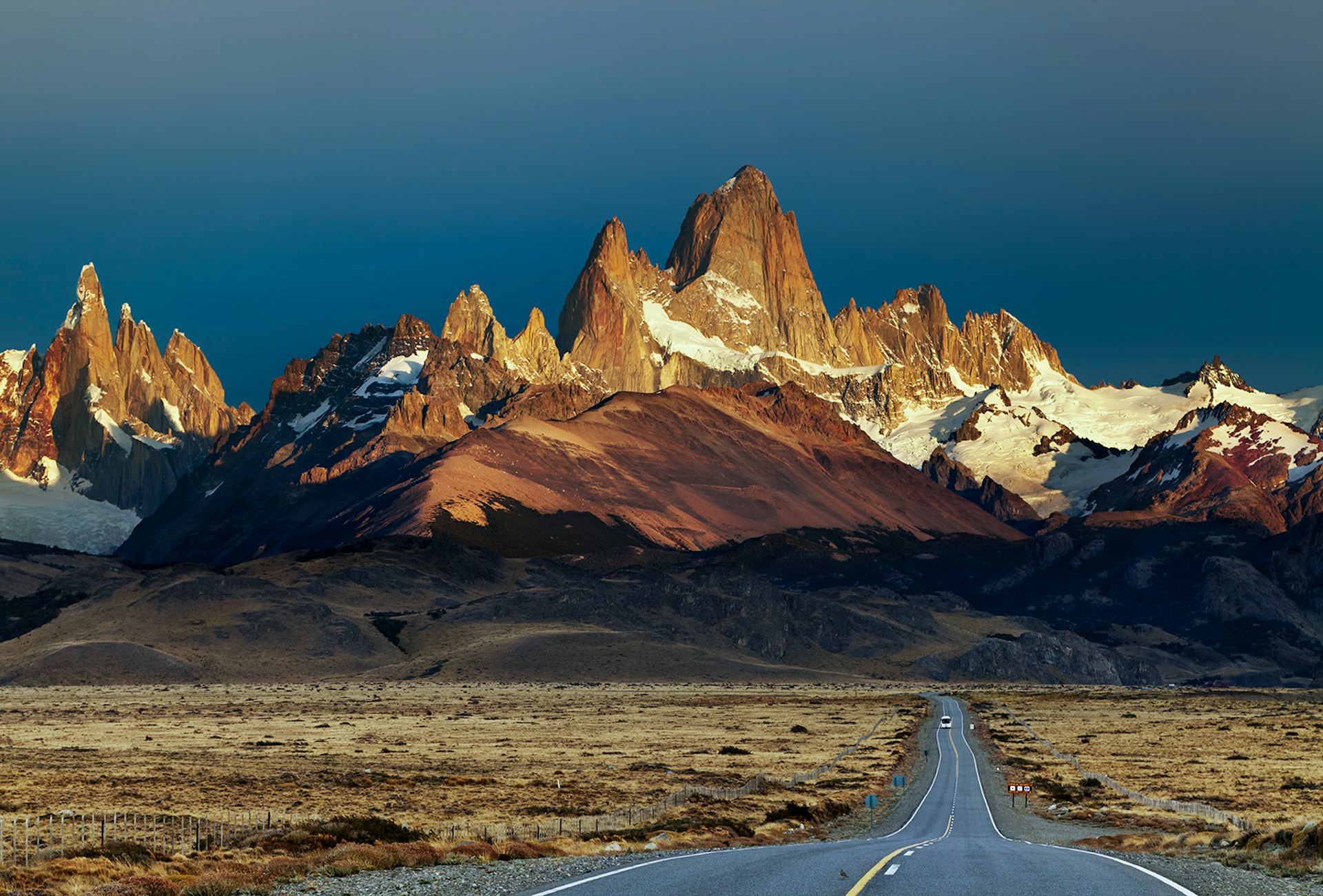 A road extends through scraggly grasslands towards the towering peaks of El Chalten at sunset. Patagonia, Argentina.