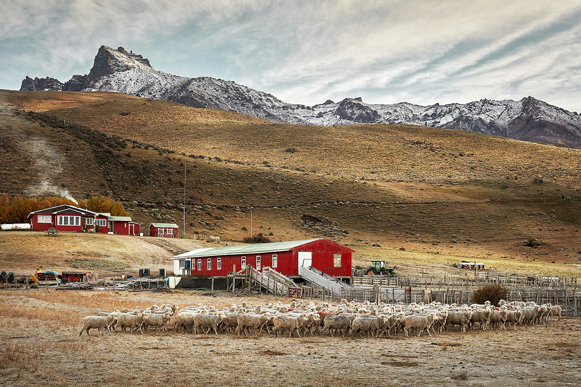 Two red buildings sit on grasslands at the food of the mountains in Patagonia. Sheep cluster in the foreground.