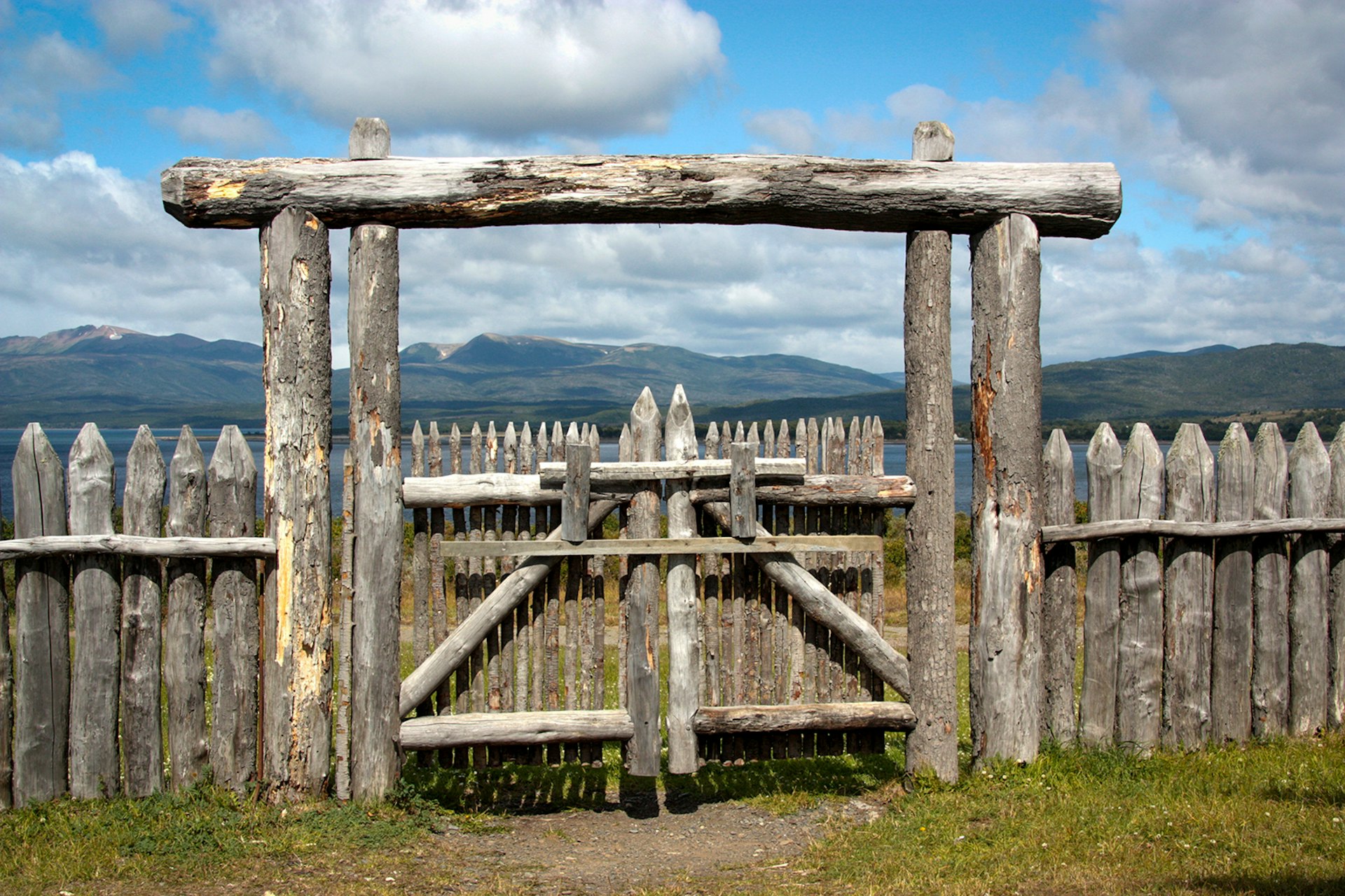 A gate constructed from pointed wooden poles stands facing water and mountains in the distance at Fuerto Bulnes. Patagonia, Chile.