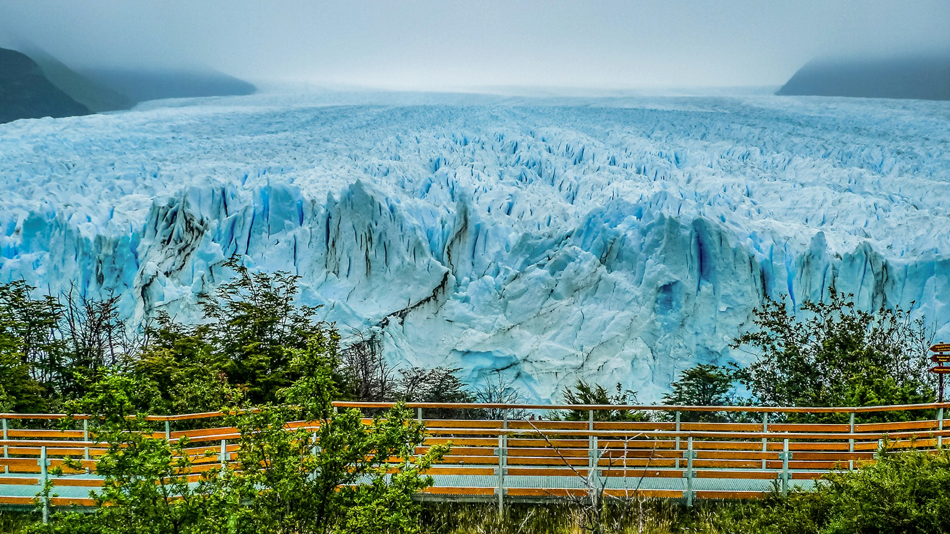 A waking bridge stretches along the front of the sheer, light blue face of Perito Moreno Glacier. Mist cloaks the hills to either side. Patagonia, Argentina.