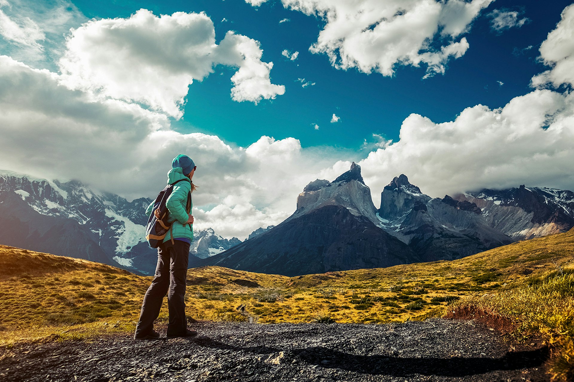A woman wearing a teal jacket walks towards mountain peaks in Torres del Paine National Park. Patagonia, Chile.