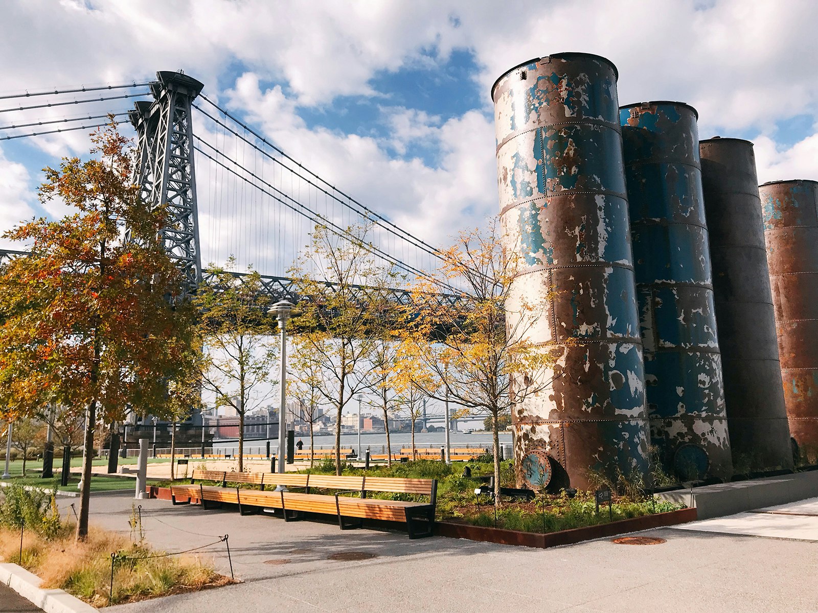 The old towers of the Domino Sugar Plant rise over a new park with the Williamsburg bridge in the background and a few trees with the leaves turning for fall on a sunny day