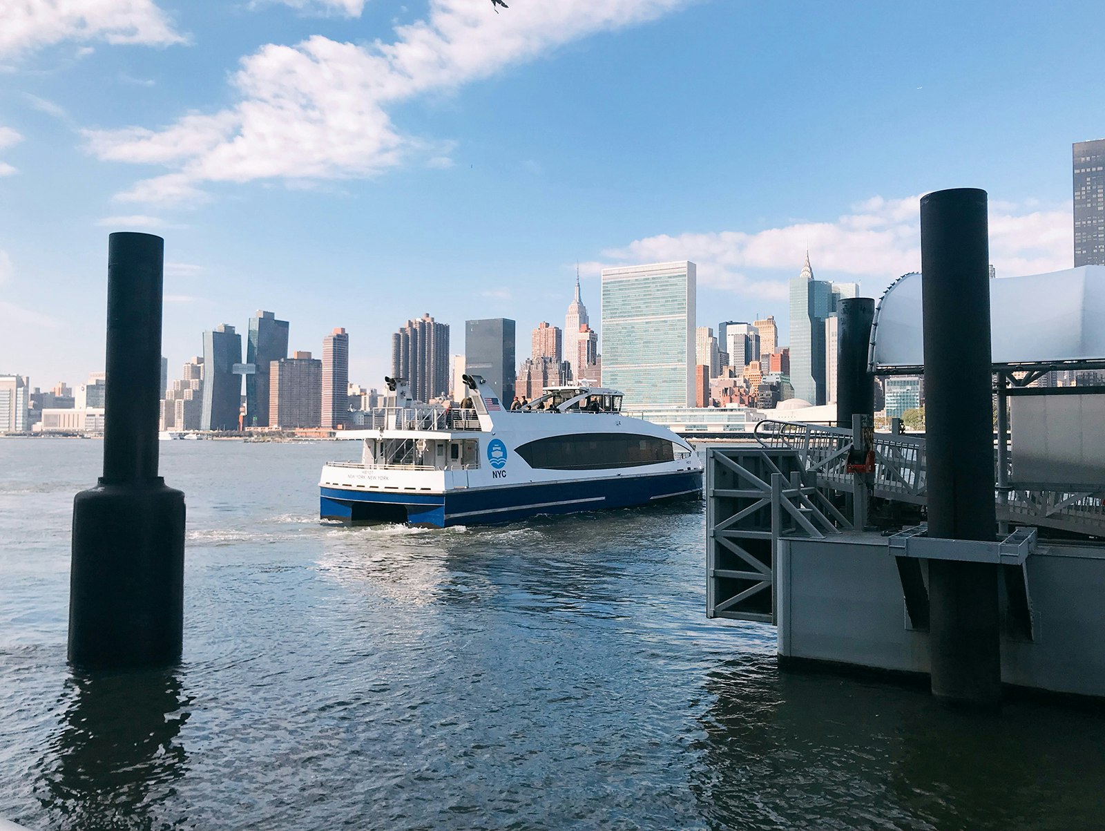 The NYC ferry floats in the harbor near Queens with skyscrapers in the background on a sunny fall day