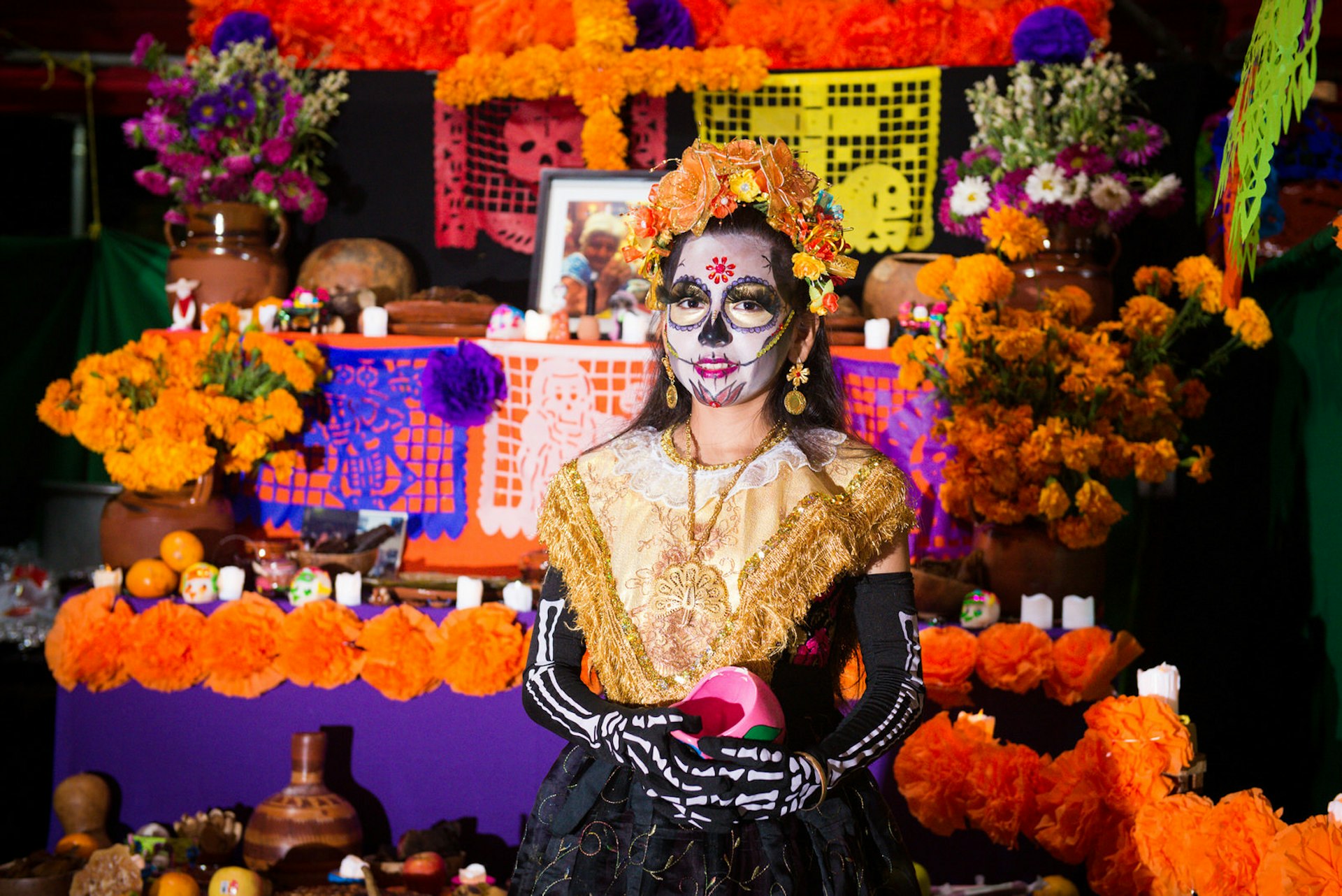Girl in skeleton costume and face paint stands in front of an altar for the Day of the Dead festival in Mexico