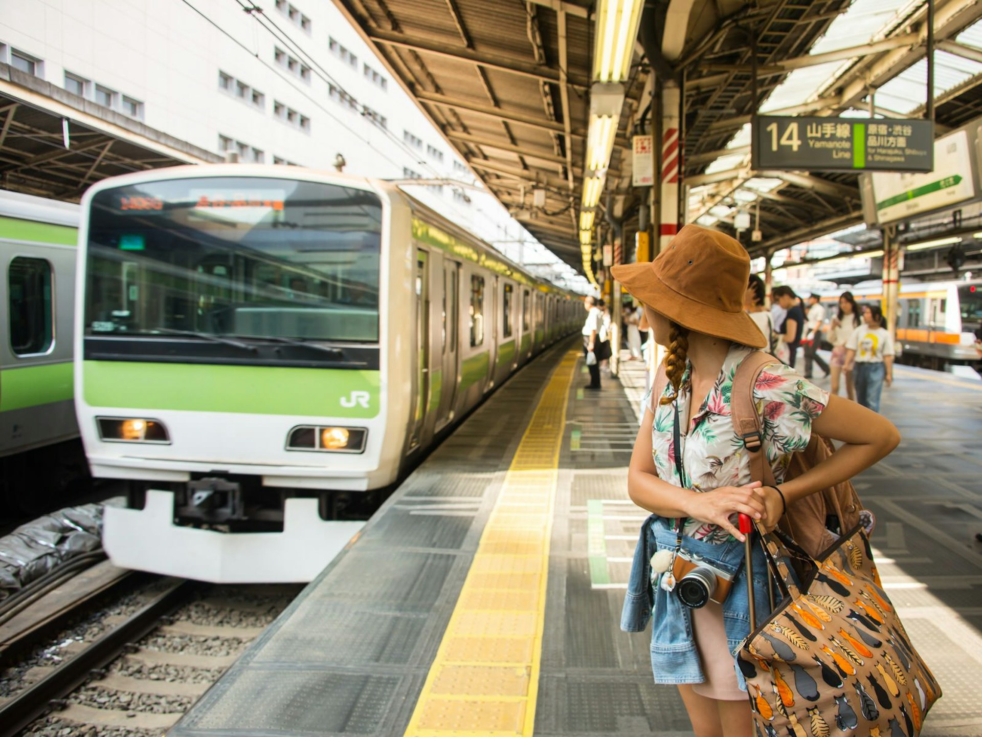 A Yamanote Line train pulls up to a station platform as a tourist with bag and camera waits to board © Vassamon Anansukkasem / Shutterstock