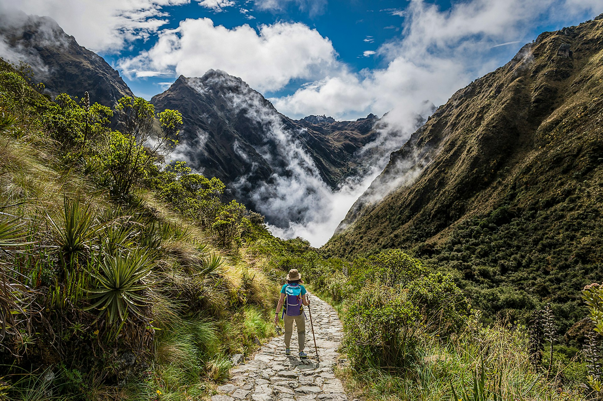 A woman in a sunhat walks down a stone path between two mountains on the Inca trail