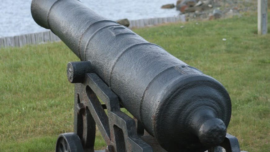 On an overcast and foggy day, an old cannon mounted on wheels is pointed out into Conception Bay with the town and church in the background in the early 1610 settlement of Cupids, Newfoundland.