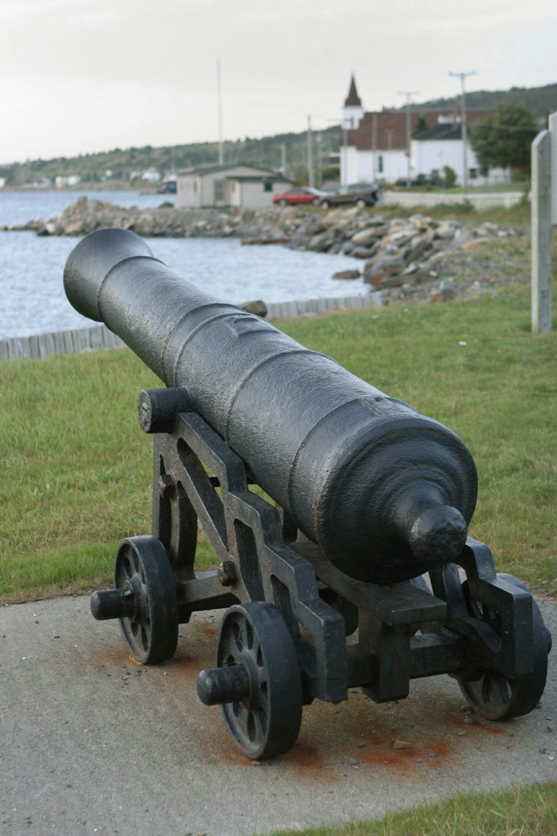 On an overcast and foggy day, an old cannon mounted on wheels is pointed out into Conception Bay with the town and church in the background in the early 1610 settlement of Cupids, Newfoundland.