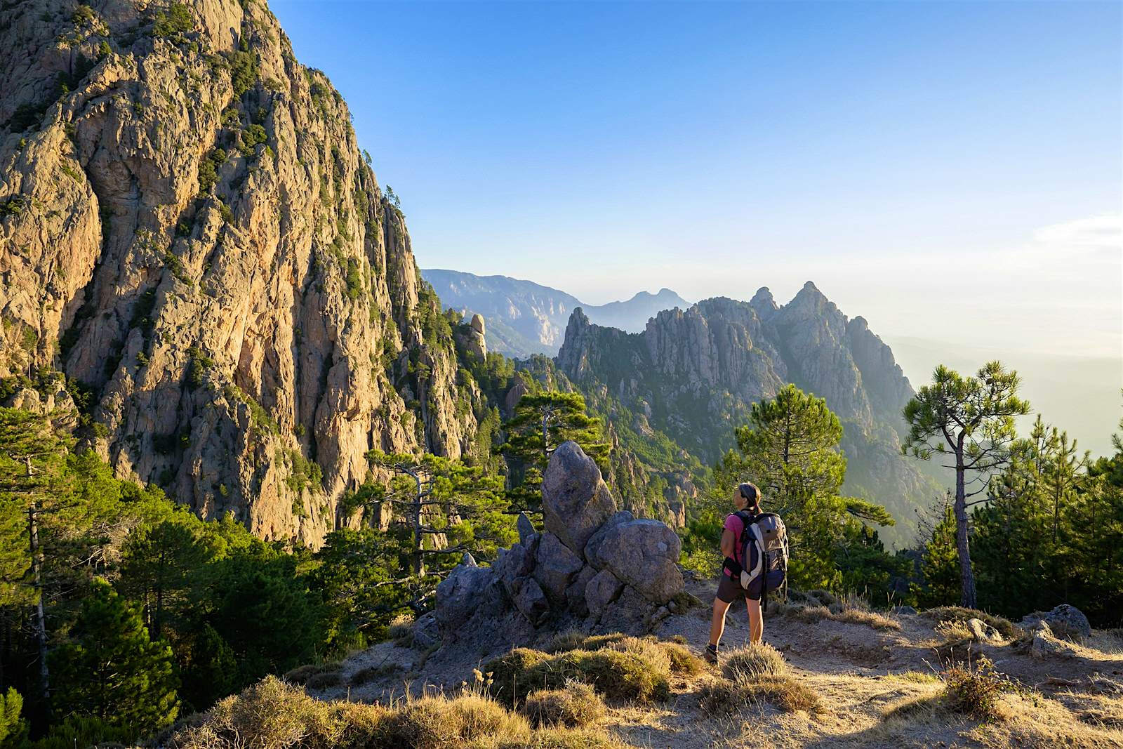 A woman with a backpack stands on a hilltop along the GR20 trail with rocky mountain peaks visible beyond her