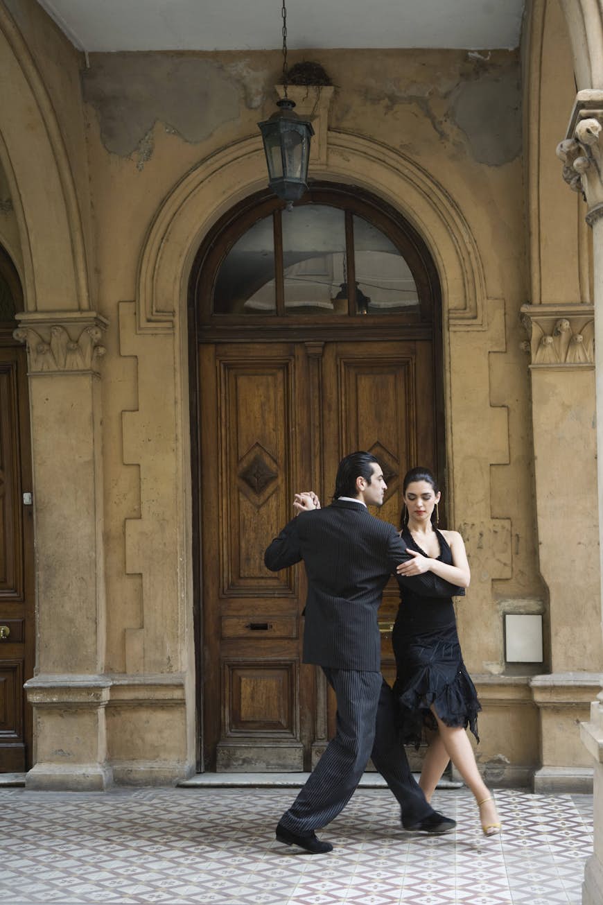 A couple, dressed all in black, dance the tango on a tiled floor in front of a large wooden door, Buenos Aires