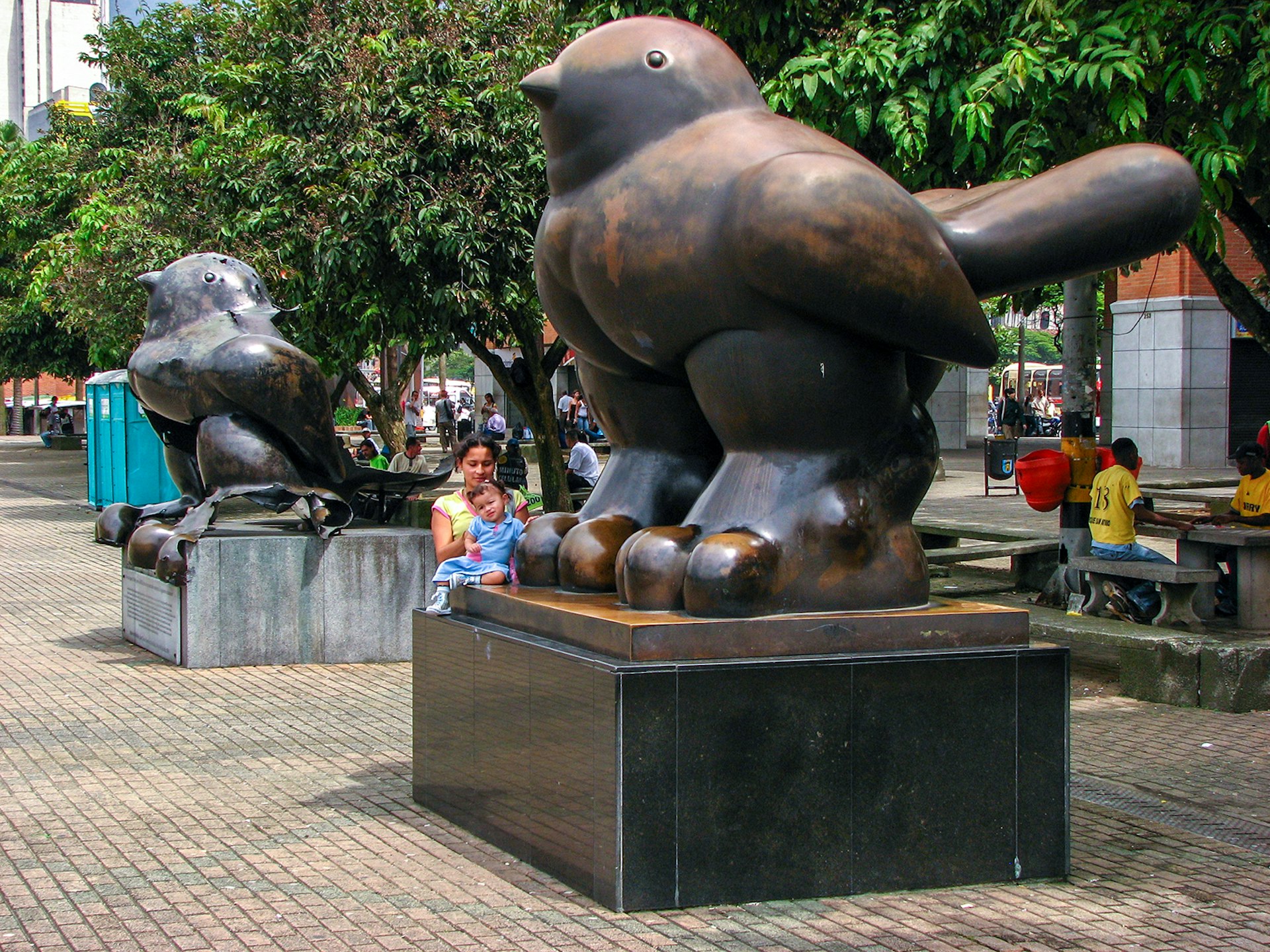Two bronze Botero statues in a park, with a woman and a baby sitting on one