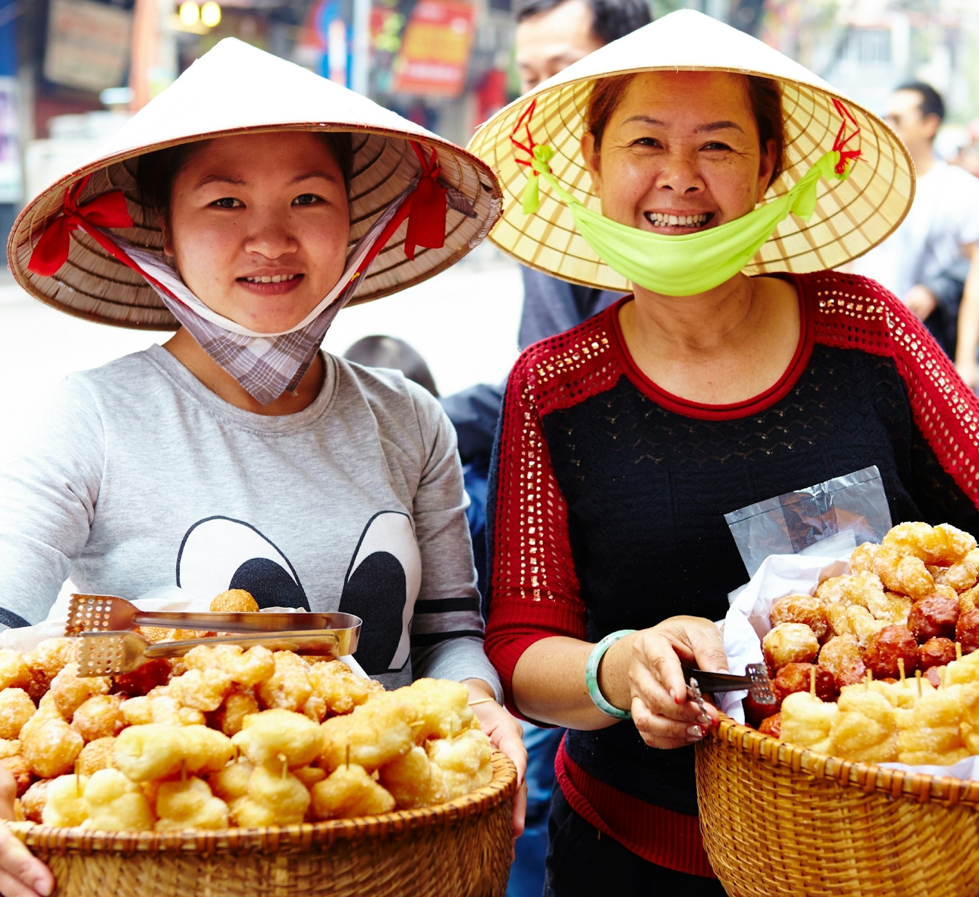 Two women pose with baskets of street food in Hanoi. Both women are wearing conical hats, which are common in the country. 