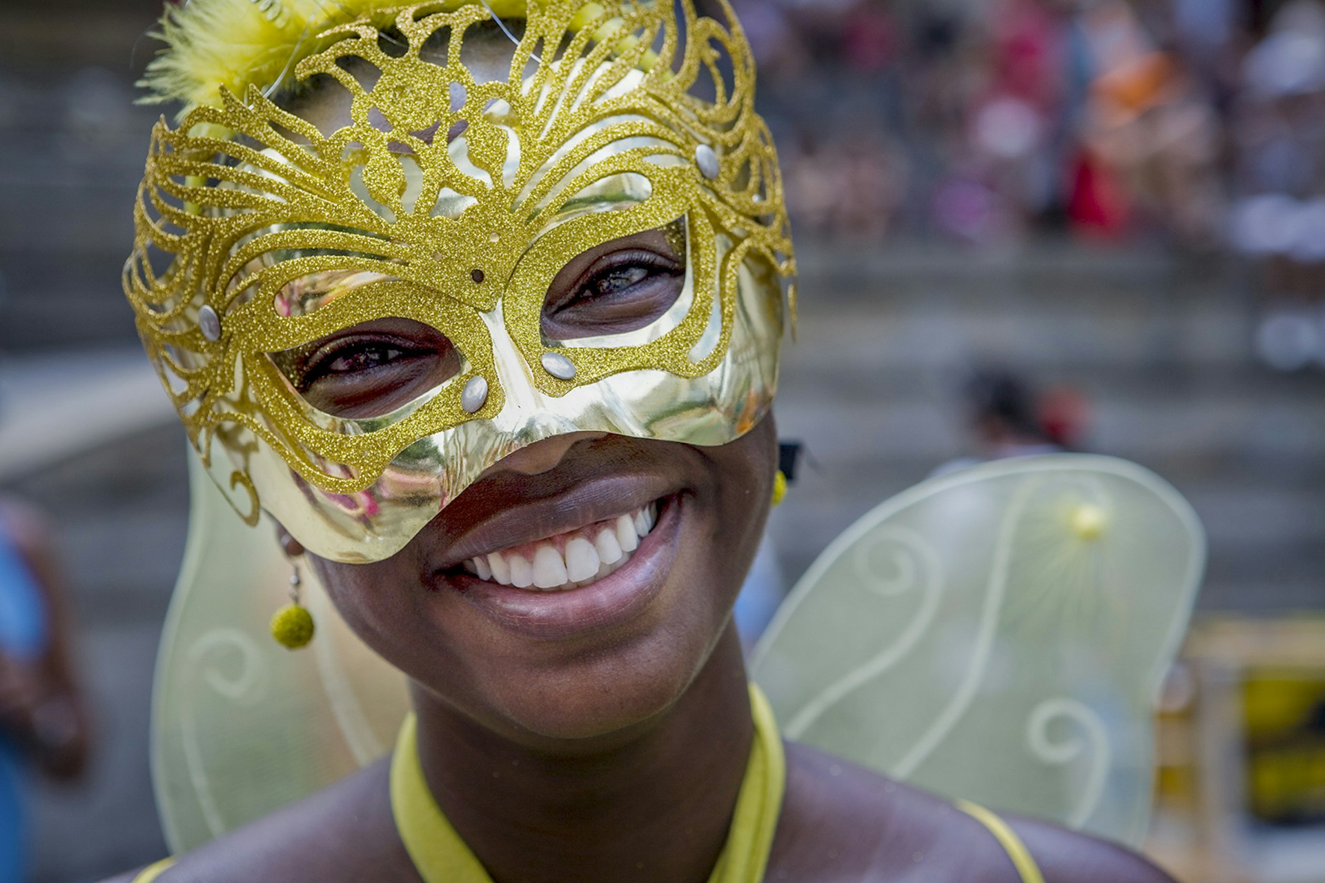 A woman wearing a gold masquerade mask and fairy wings smiles at the camera