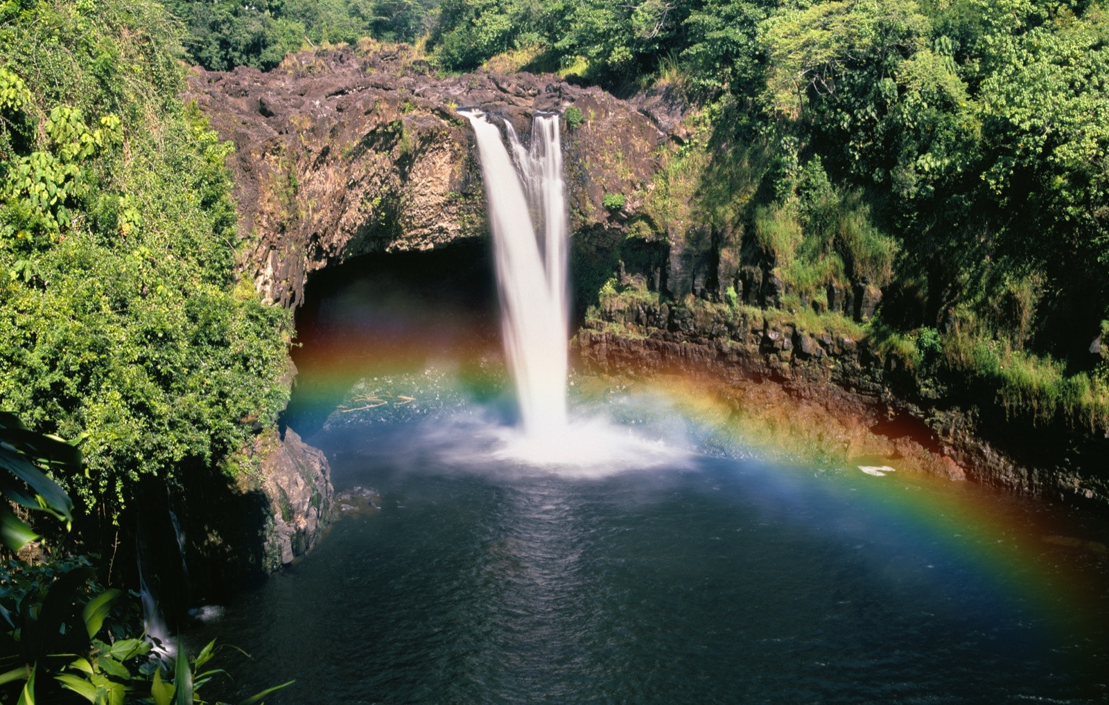 A rainbow encircles a waterfall emptying into a large pool of water surrounded by lush green trees. Hawaii's Big Island has a full tray of offerings. 