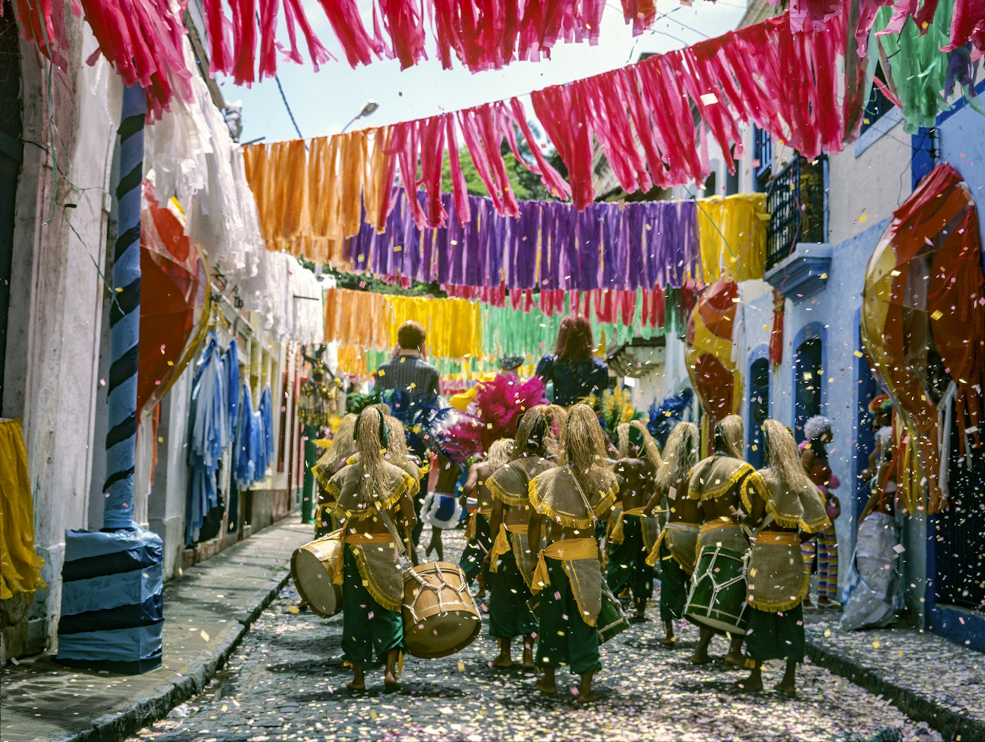 People wearing green and gold skirts and headdresses walk beneath colorful paper fringe hanging over a historic street