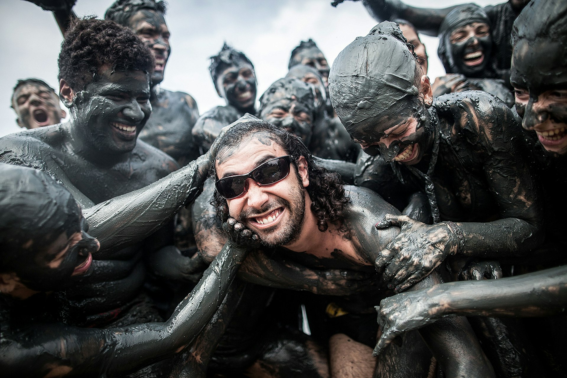 A man in sunglasses gets smeared with dark mud by dozens of carnival goers