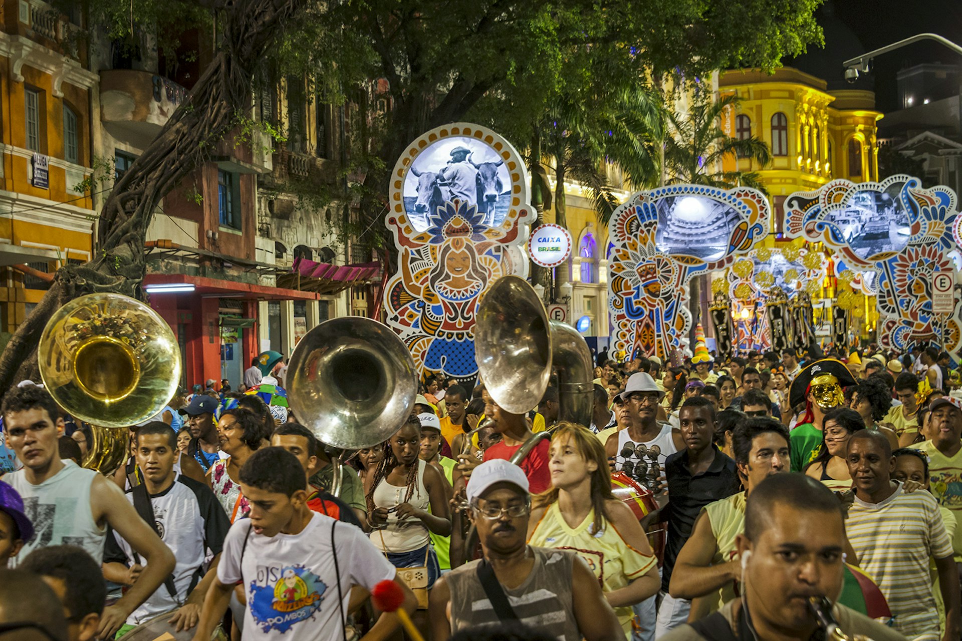 A group of people led by those holding tubas walking down a colorful street, holding thematic parade signs